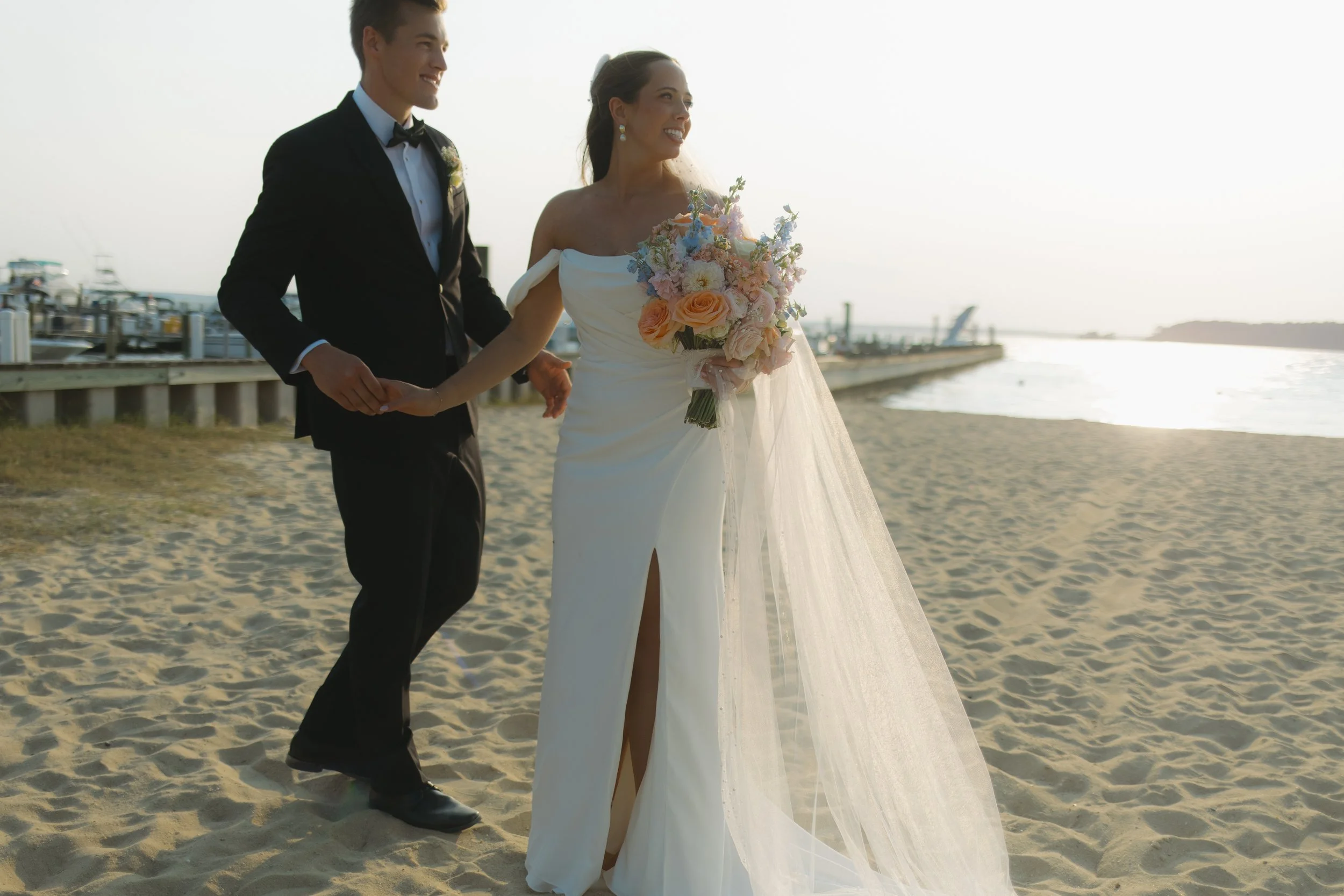 A bride and groom walk on the beach at sunset, holding hands. The bride wears a white wedding gown with a slit, and holds a bouquet of pastel-colored flowers. The groom wears a black tuxedo with a bow tie.
