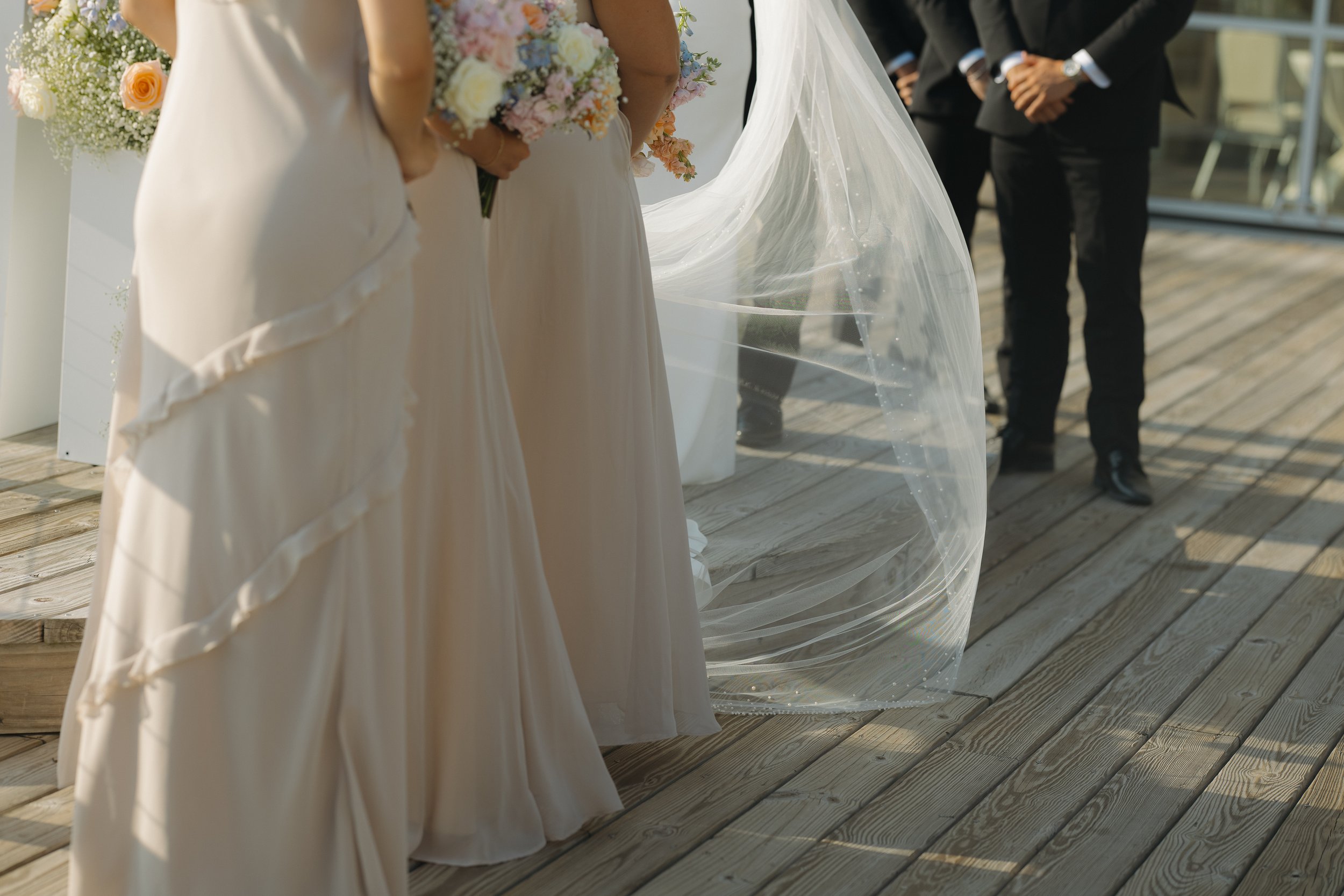 Bridal party standing on a wooden deck with bridesmaids in beige dresses holding bouquets and a bride in a white wedding gown with a long veil, alongside groomsmen in black suits.