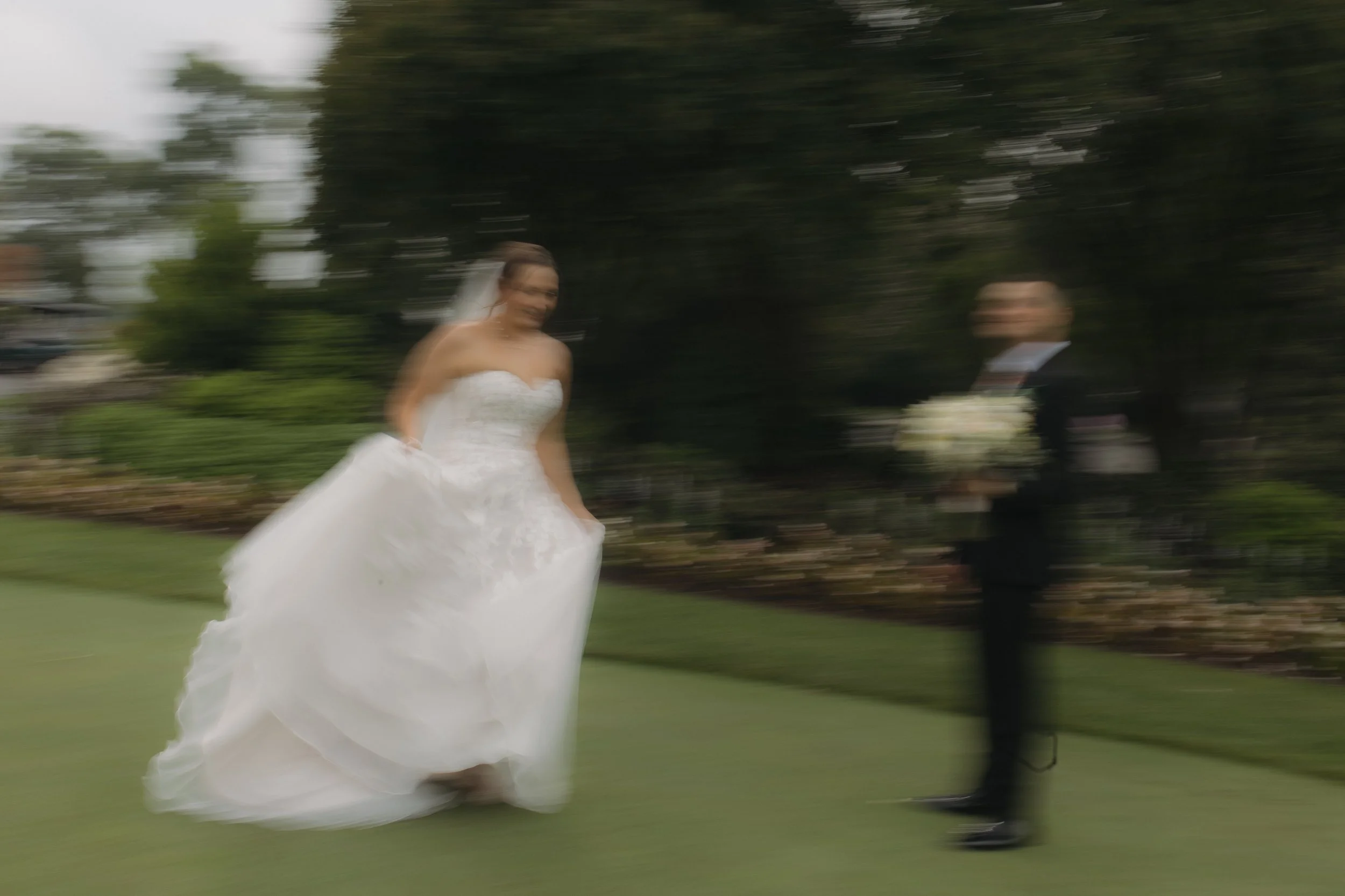 Blurry photo of a wedding bride in a white gown and veil holding the skirt of her dress and a groom in a black tuxedo holding a bouquet, standing on a grassy area with trees in the background.