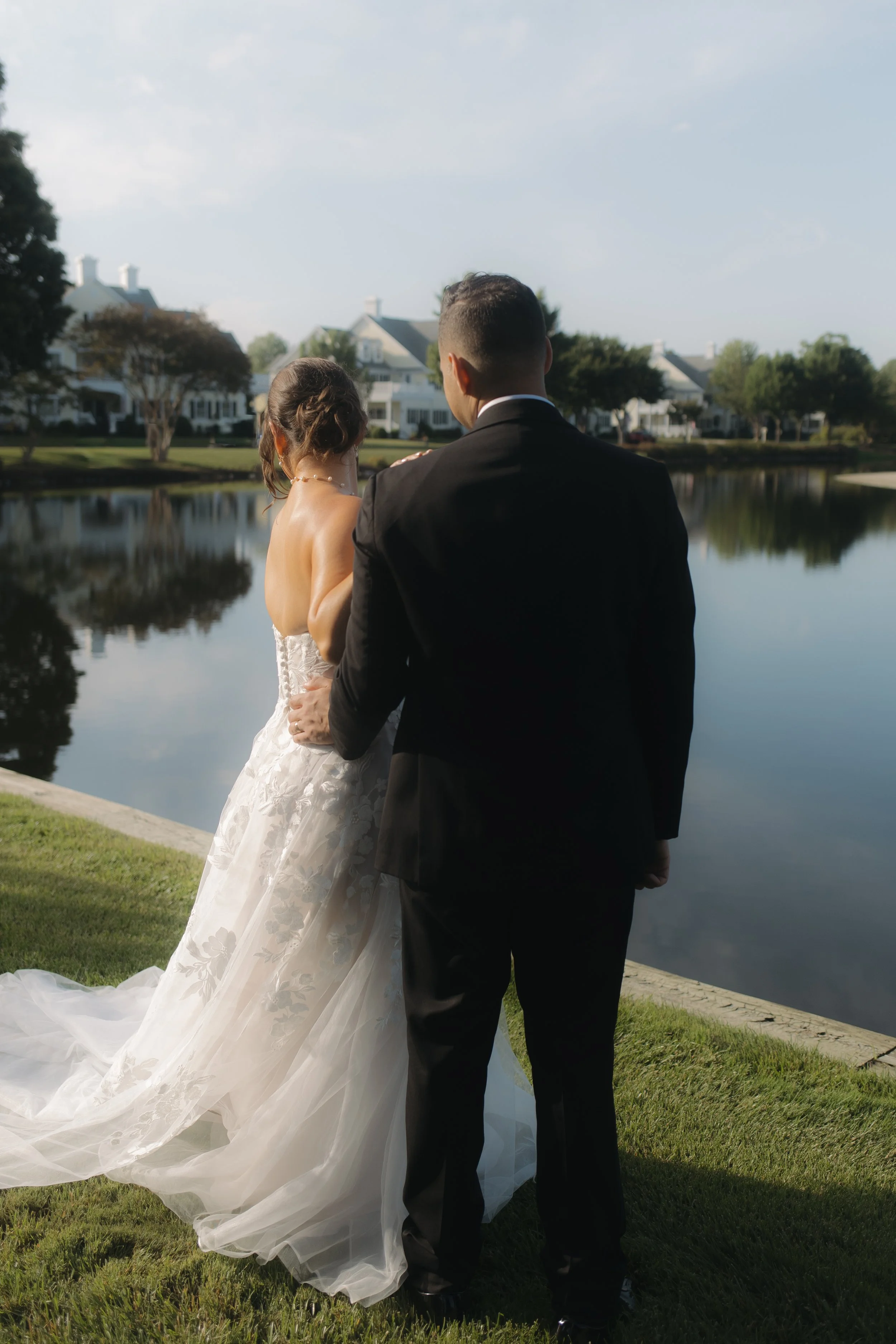 A bride and groom standing by a lake on their wedding day, with houses and trees in the background.