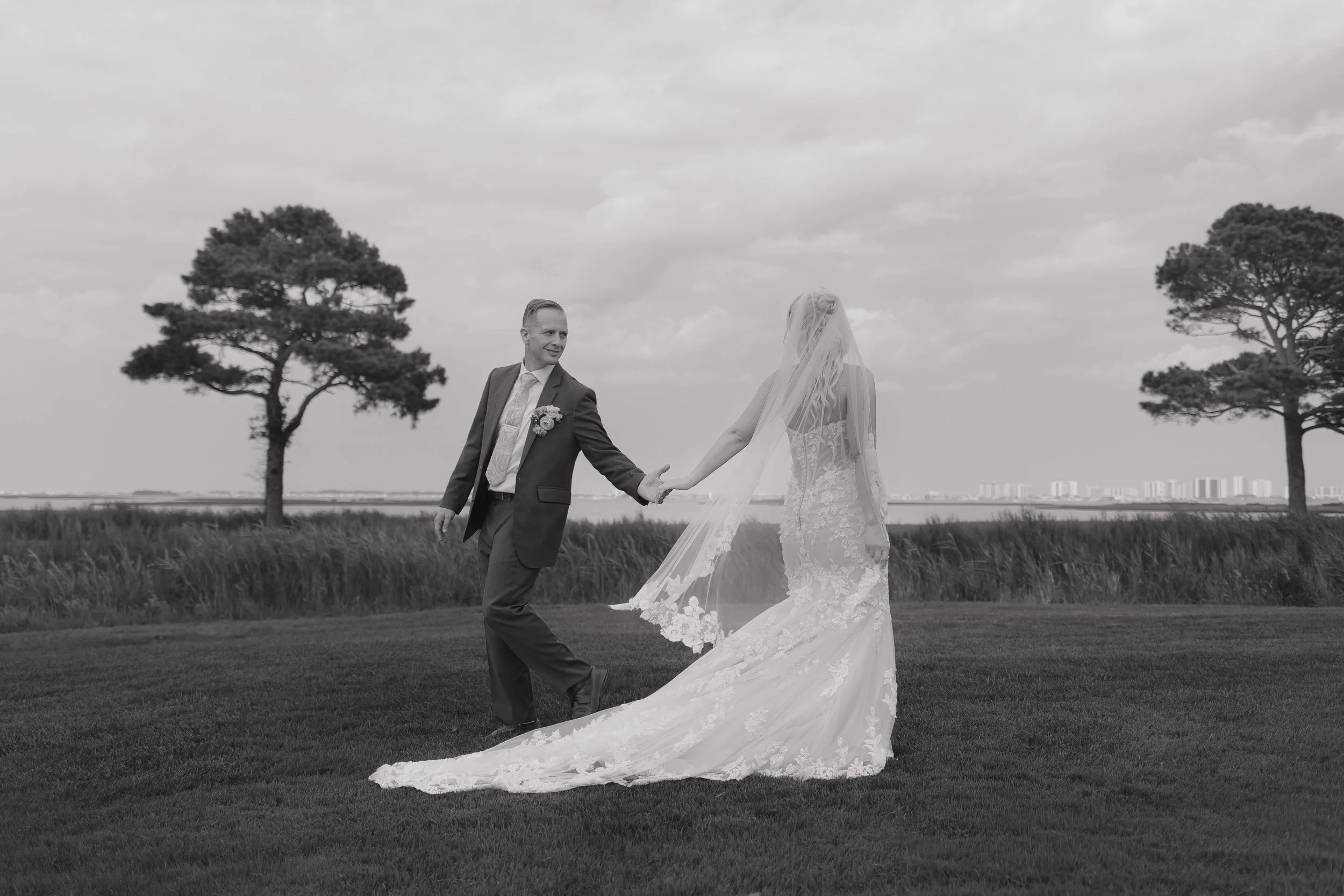 A black and white photograph of a bride and groom on a grassy field, holding hands, with trees and a cityscape in the background. The bride wears a lace wedding gown with a long train and veil, and the groom wears a suit with a boutonniere.