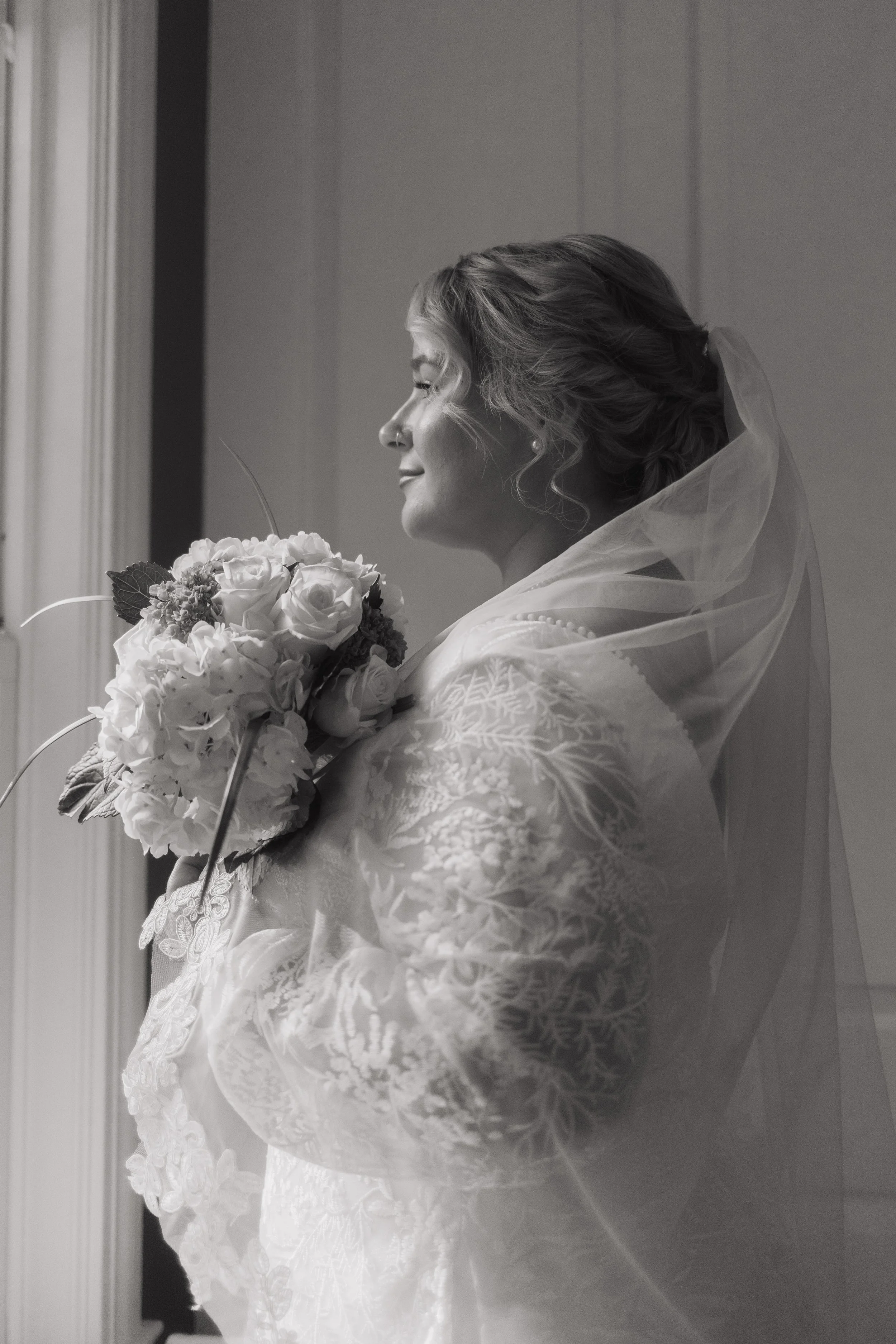 A black and white photo of a bride holding a bouquet of flowers, wearing a wedding dress and veil, standing indoors near a window.