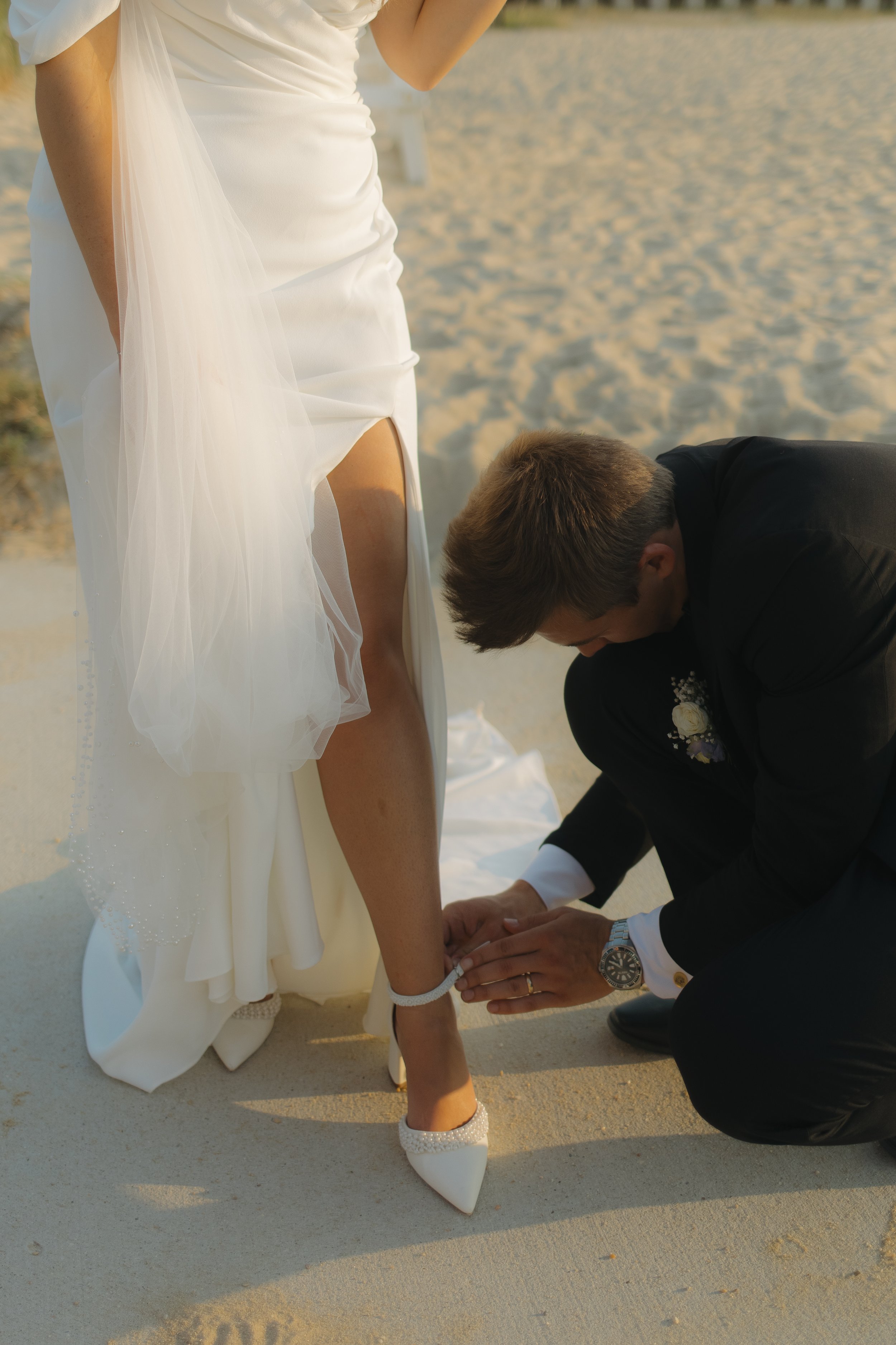 A groom helping a bride with her shoe on a sandy beach during a wedding ceremony.