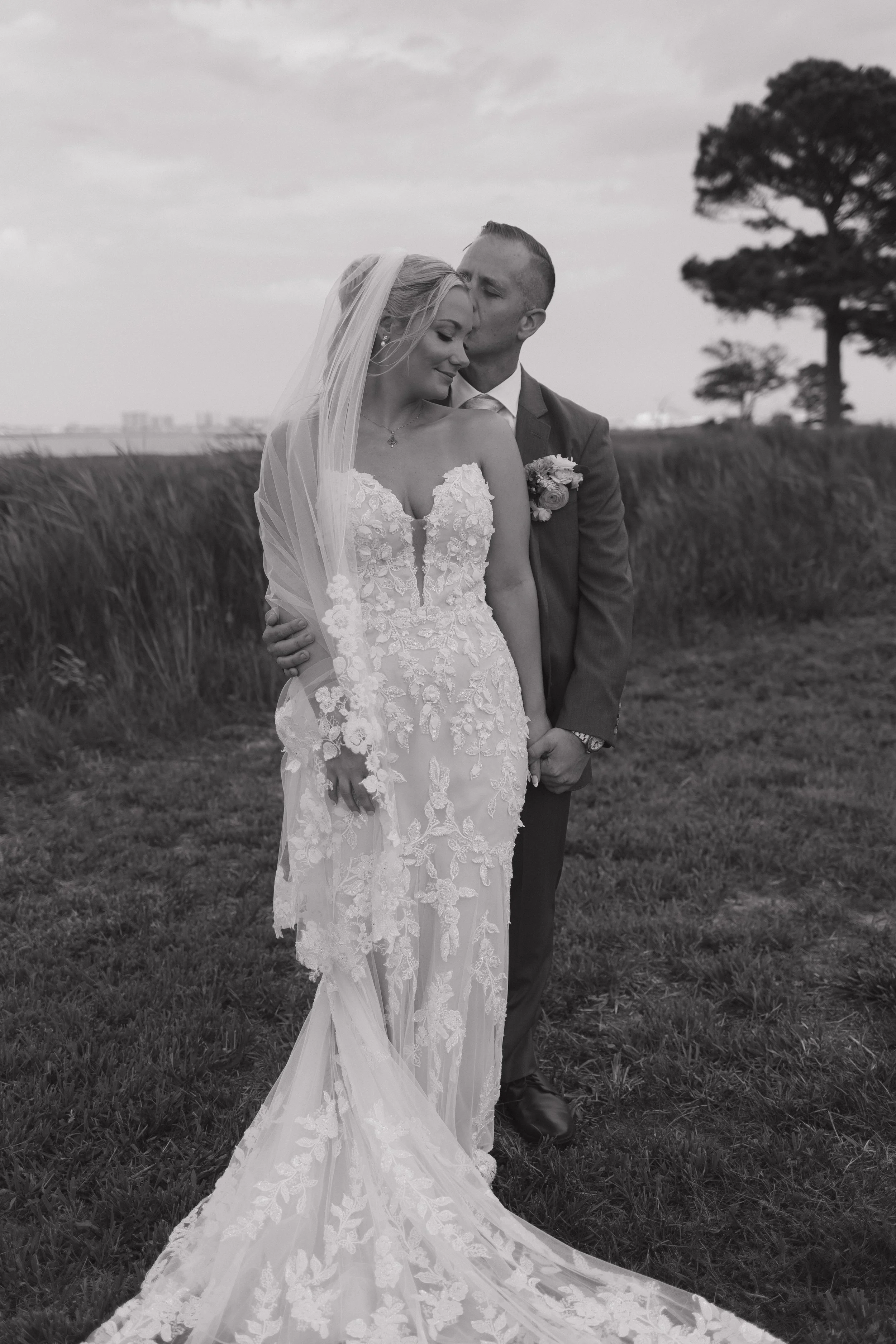 Black and white photograph of a bride and groom standing outdoors on grass, with the groom kissing the bride's forehead and holding her hand. The bride is wearing a detailed lace wedding gown with a veil, and the groom is in a suit with a boutonniere