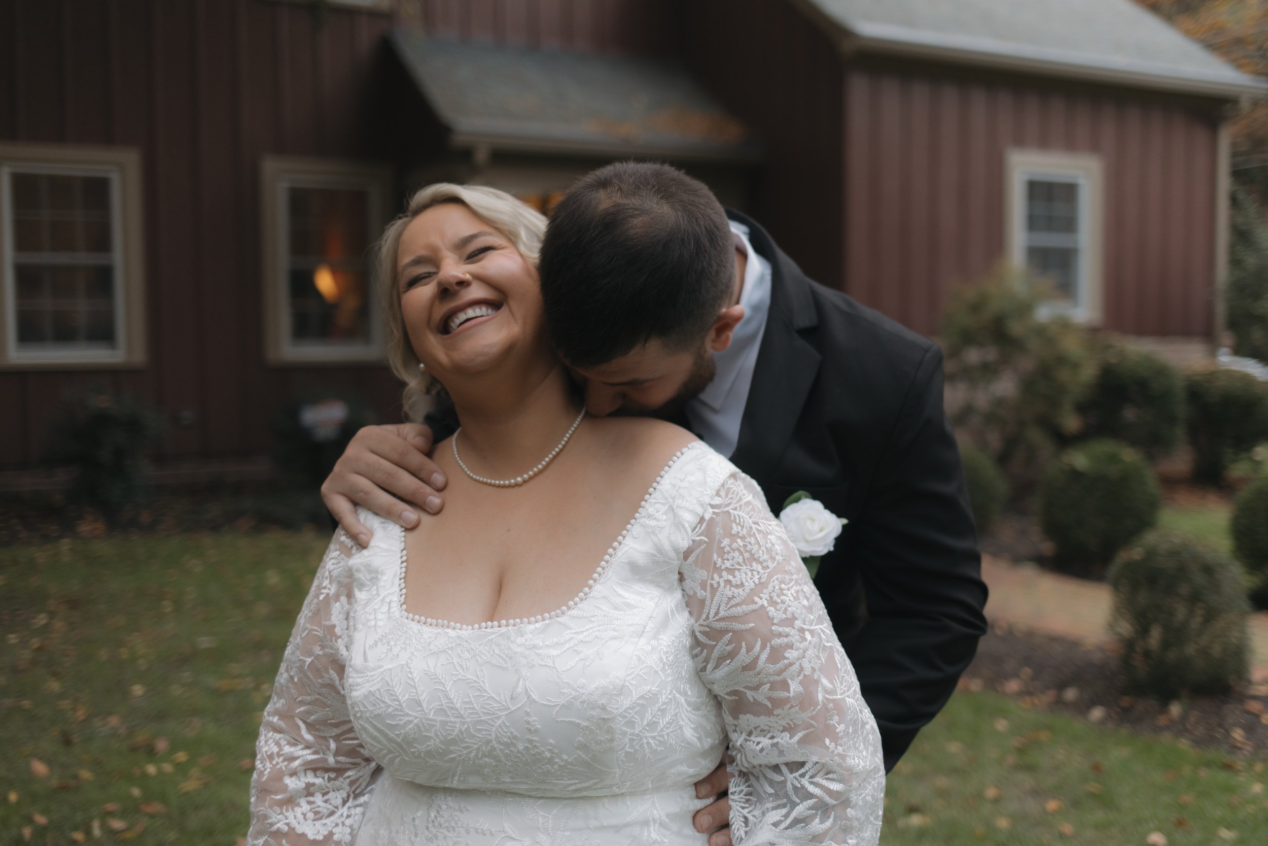 A bride with a joyful smile, wearing a white lace dress and pearl necklace, is being kissed on the neck by a groom in a black suit outside a rustic house.