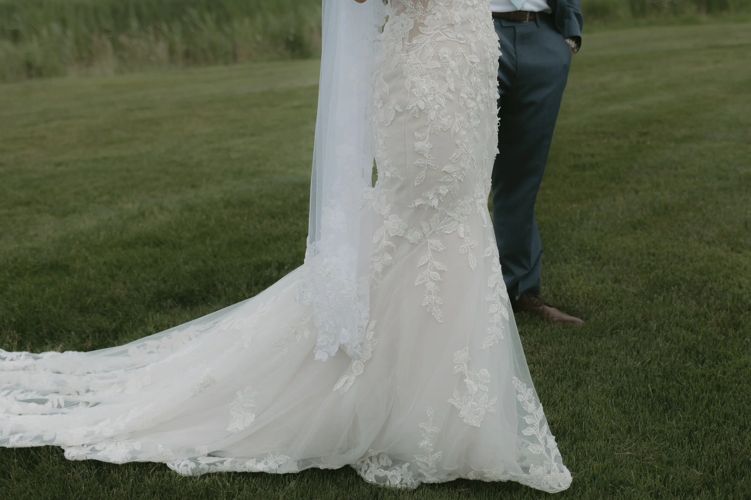Close-up of a wedding dress with lace details and a long train, standing on grass with a person in pants and shoes in the background.