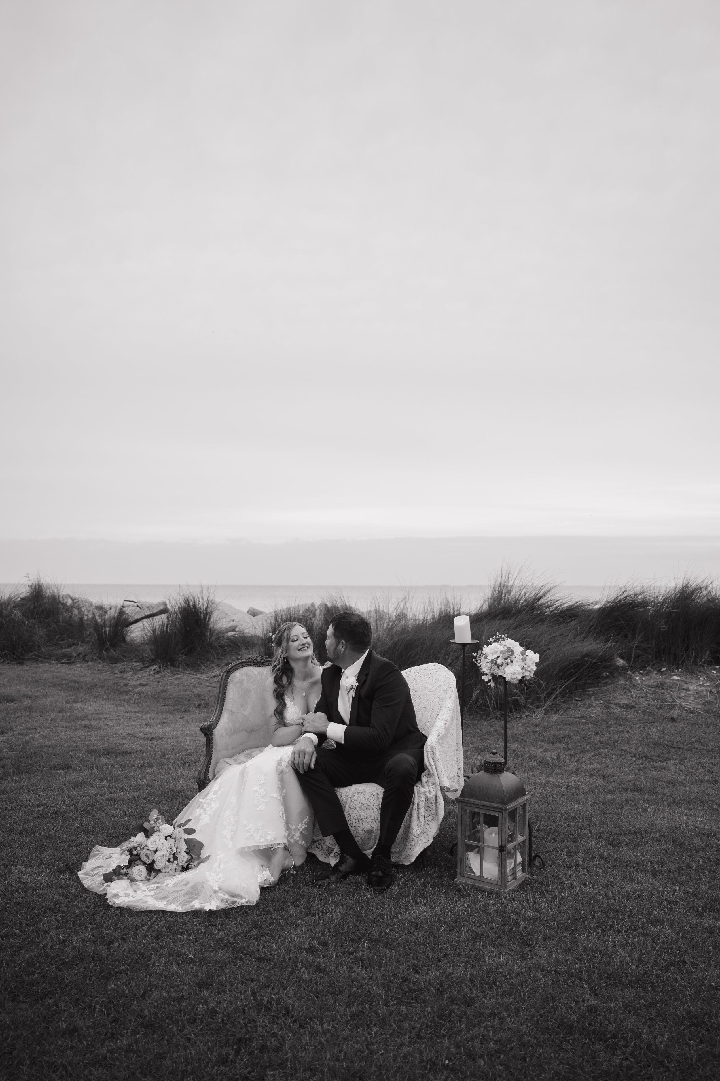 Black-and-white photo of a bride and groom sitting on a vintage sofa outdoors near the beach, sharing a moment of affection with a bouquet on the grass and candle holders nearby.
