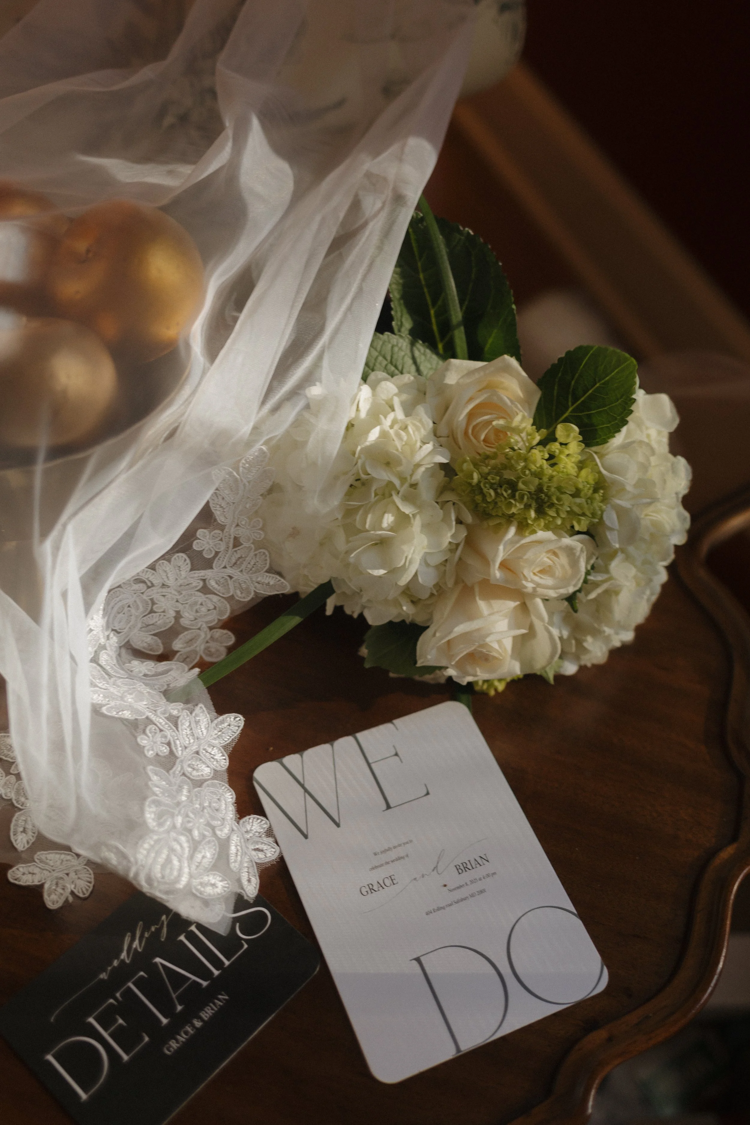Wedding bouquet of white roses and hydrangeas, a card with the words 'Welcome' and 'Grace and Brian,' a lace doily, and a black card that says 'Valbary Details Grace & Brian' on a wooden table.