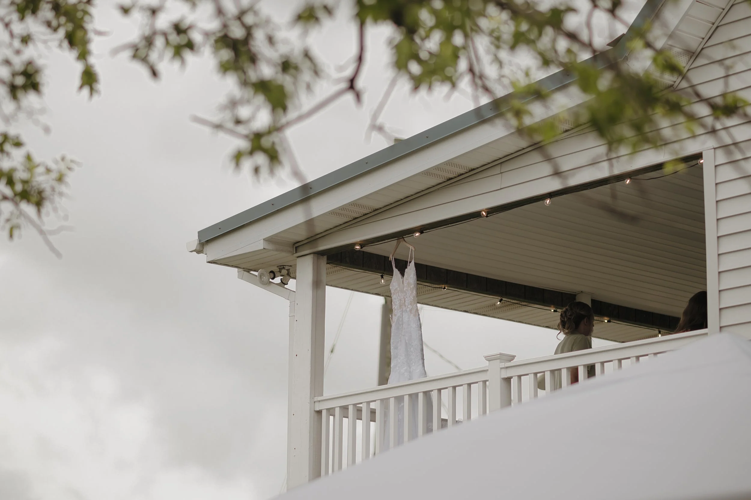 A wedding dress hanging on the ceiling of a porch, with two women standing nearby, under a roof with string lights, and the sky in the background.