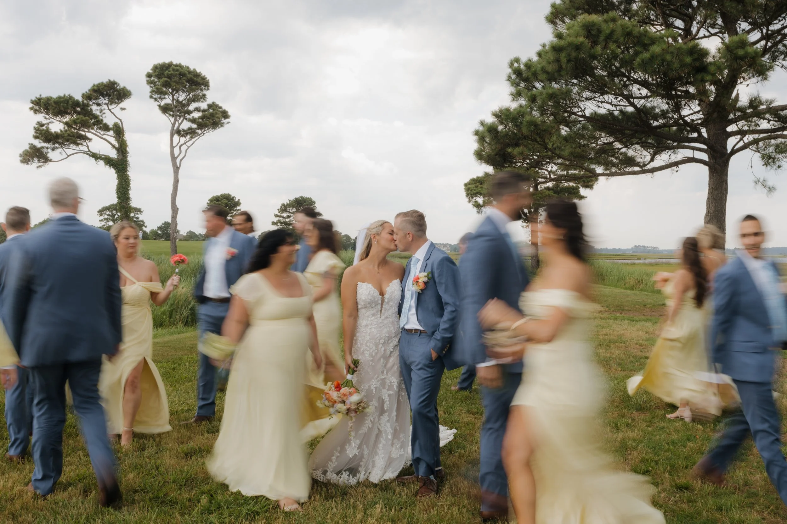 A bride and groom kissing in the center of a group of wedding guests outdoors on a grassy field with trees and cloudy sky in the background.