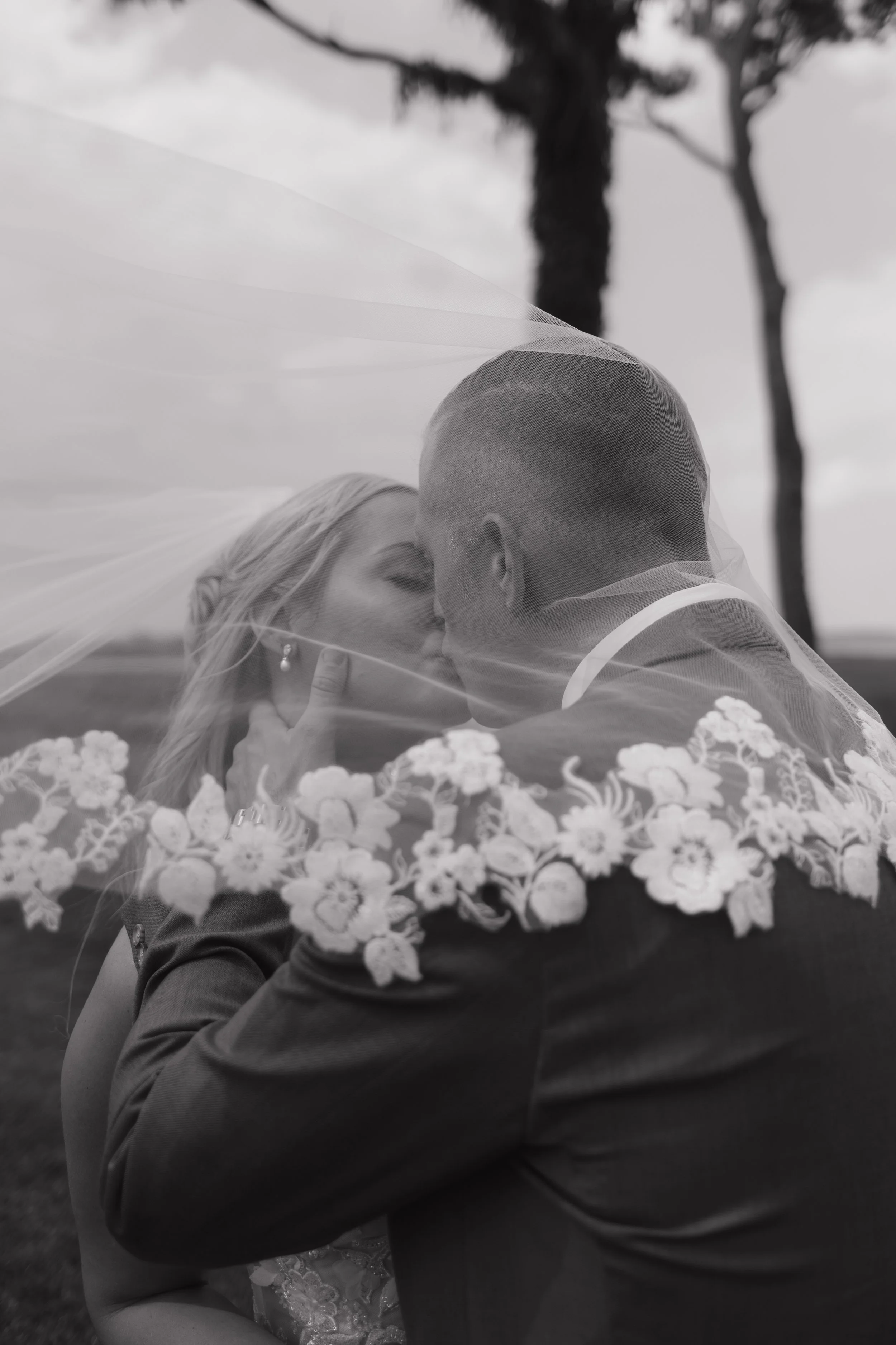 A black-and-white photo of a couple kissing under a wedding veil outdoors, with trees in the background.