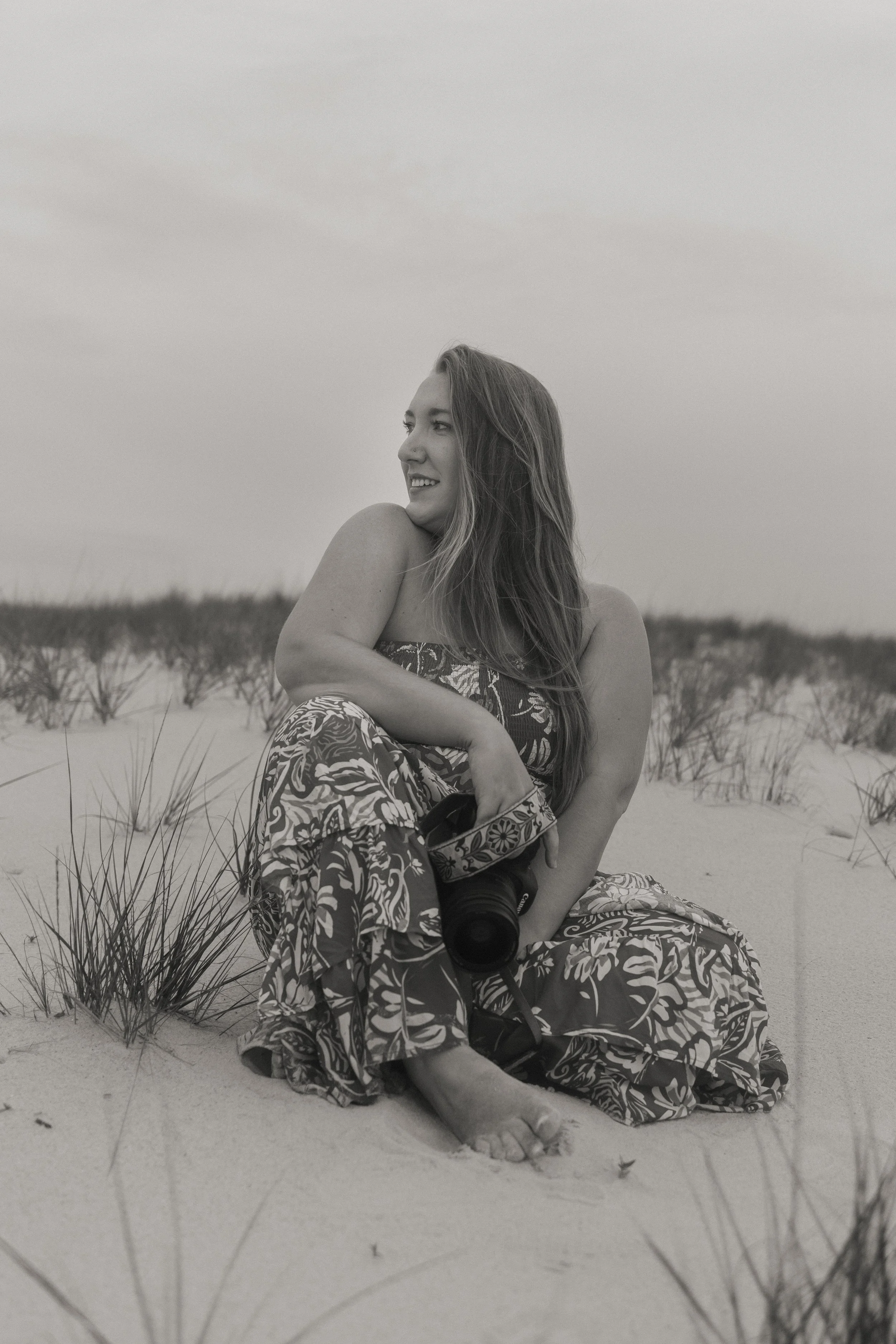 A woman sitting on the sand in a beach setting, holding a camera, smiling, with sparse vegetation around her, black and white photo.