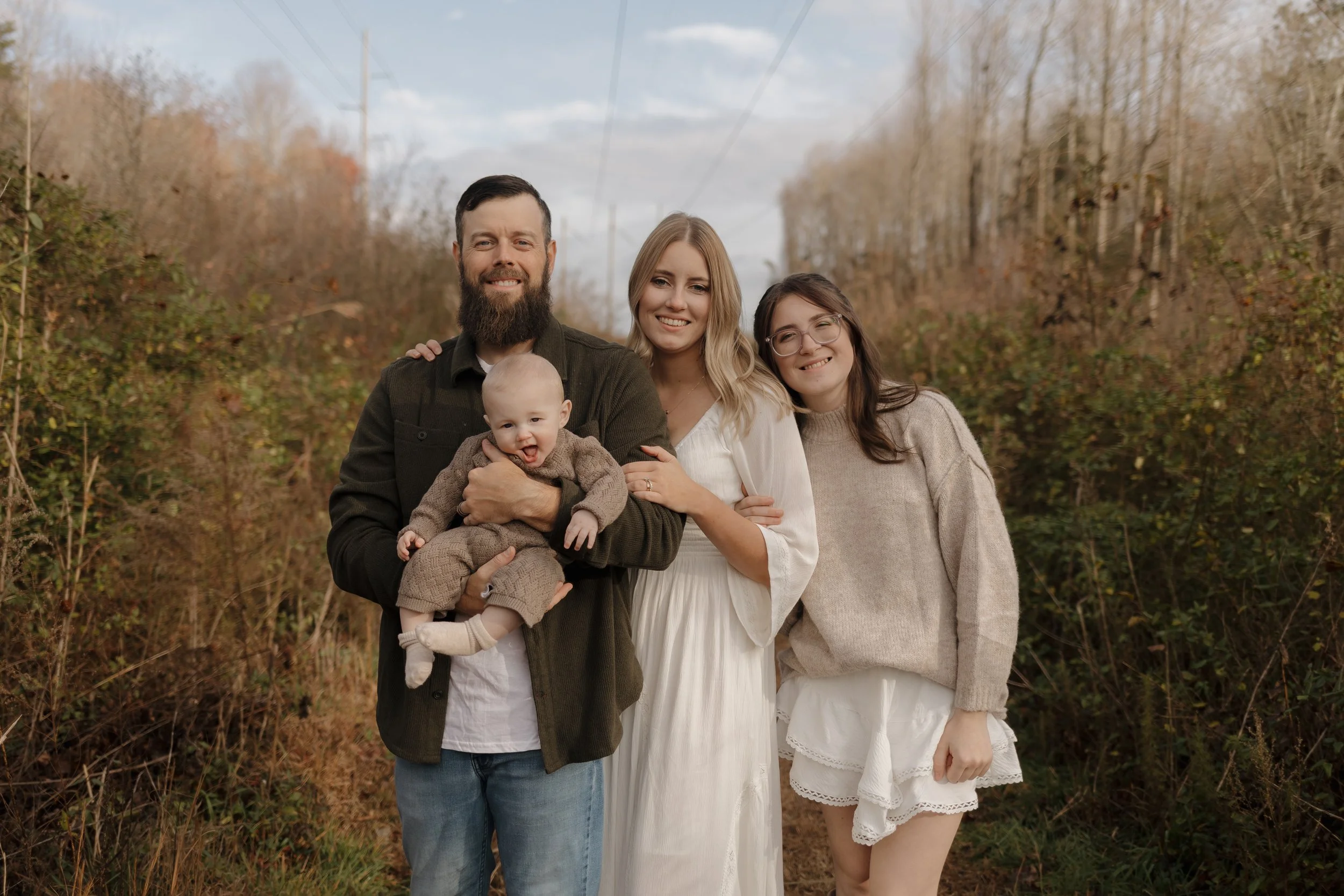 A family of four standing outdoors in a natural setting with trees and power lines in the background. The man has a beard and is holding a baby. The woman on the right is wearing glasses, and the woman in the middle is in a white dress. They are smiling and close together.
