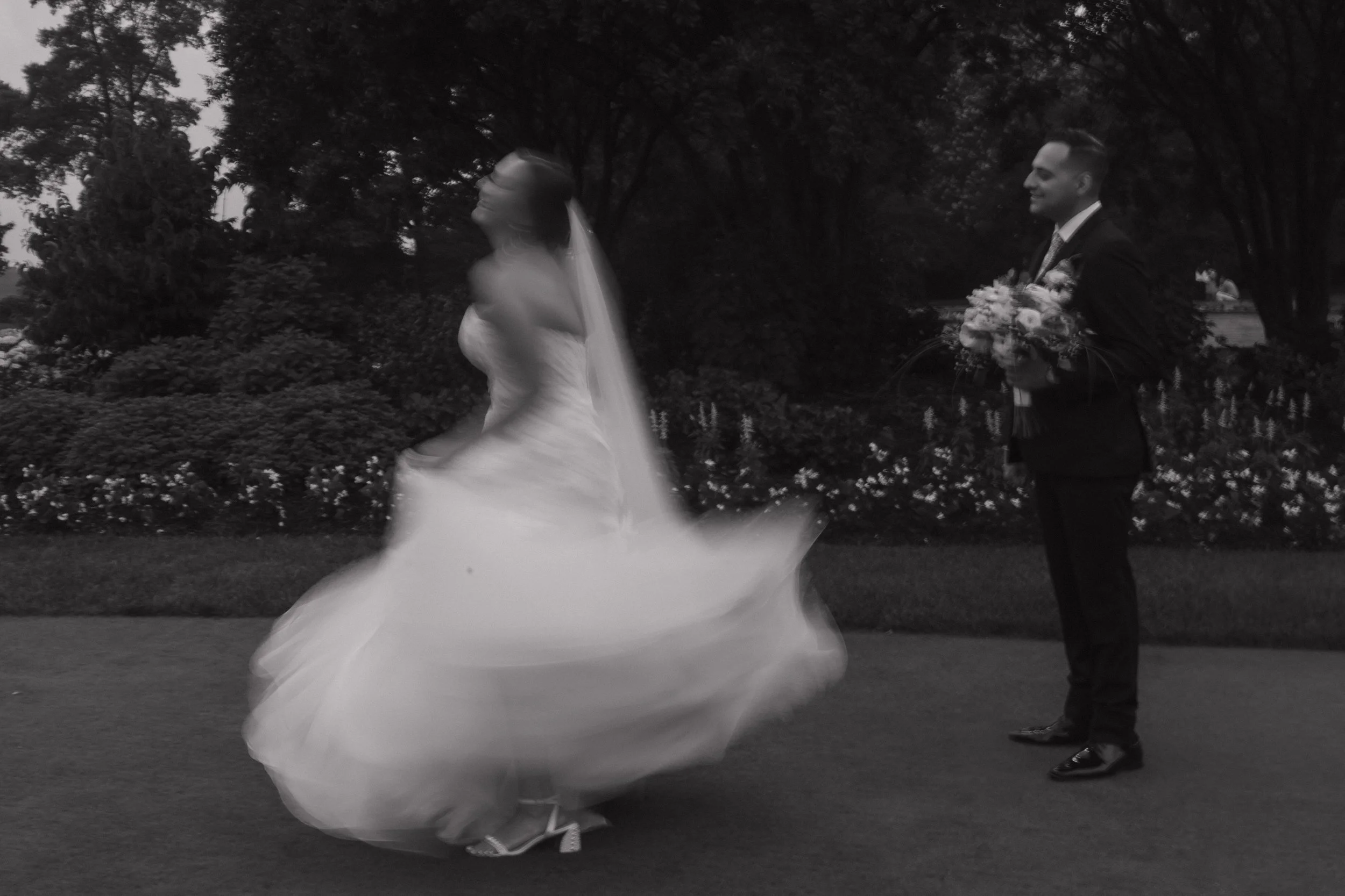 A black and white photo of a bride and groom in a garden, with the bride spinning in a wedding dress and the groom holding a bouquet, standing and smiling.