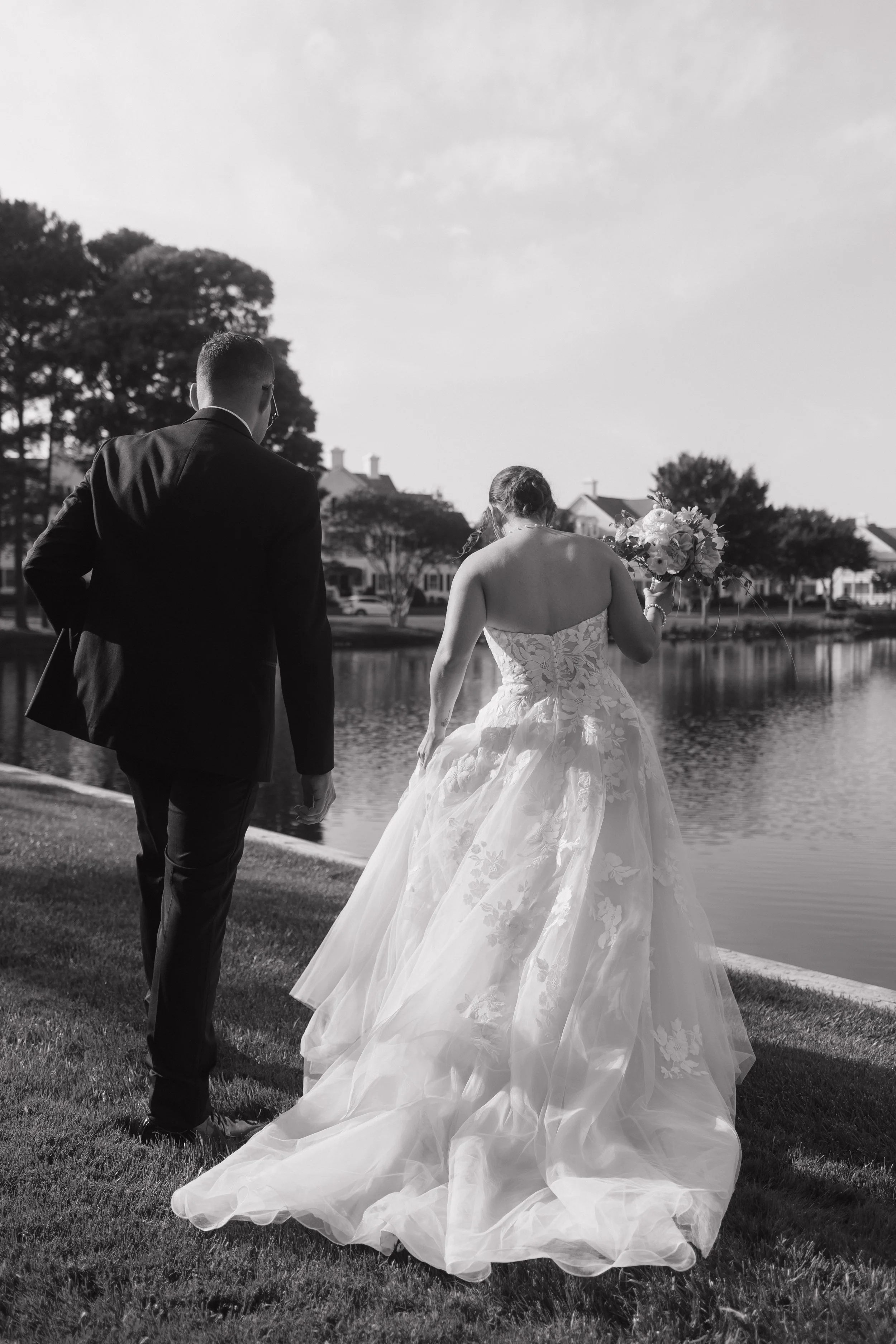 A black-and-white photo of a bride and groom walking by a lake, with the bride holding a bouquet.