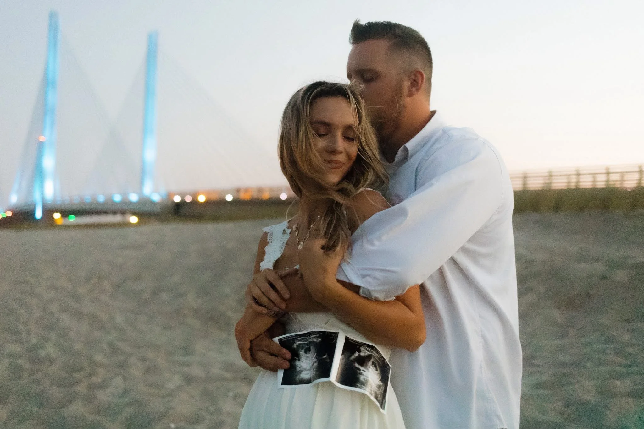 A man and woman embracing on a beach during sunset, the woman holding ultrasound images.