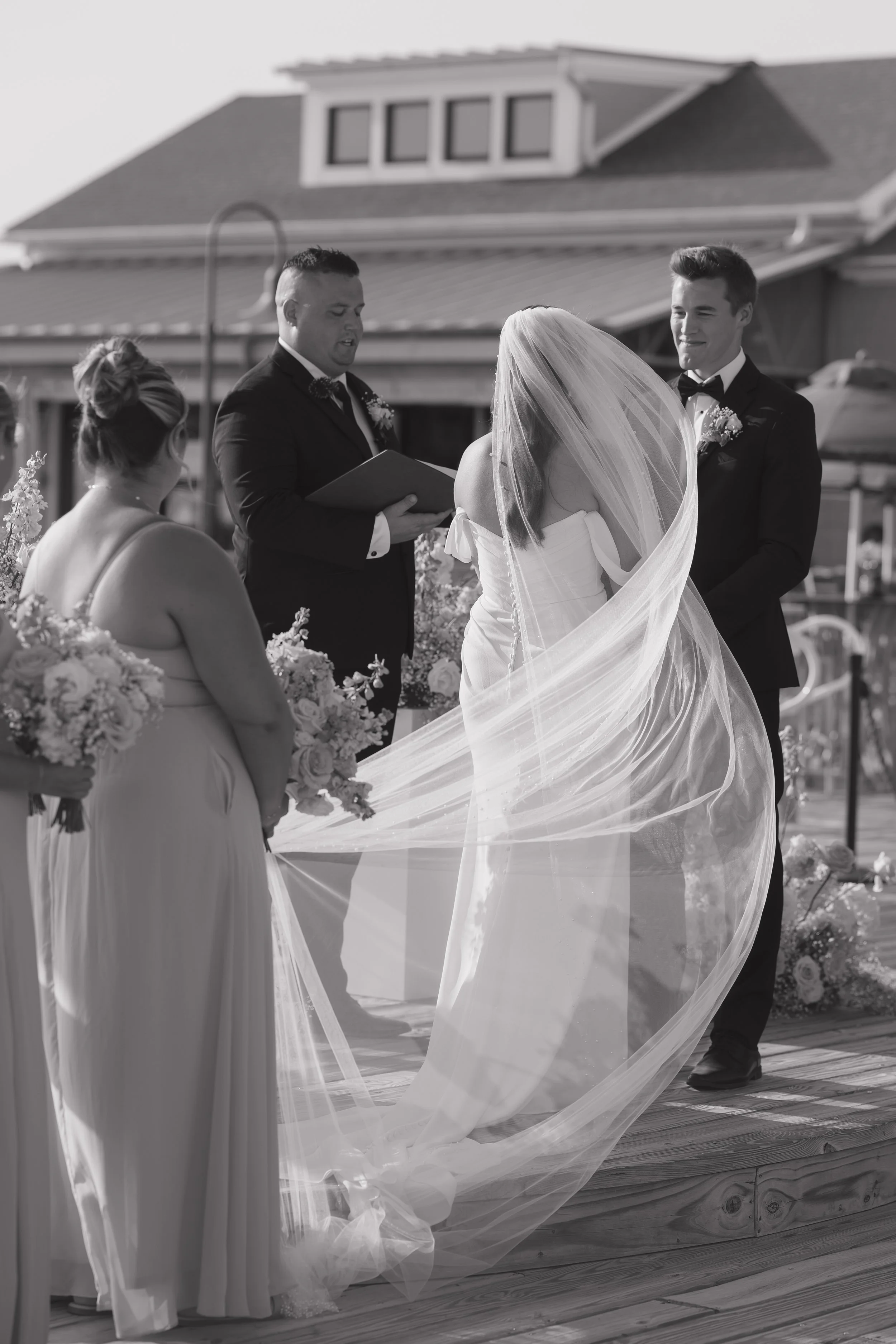 A black and white photo of a wedding ceremony outdoors, with a bride in a flowing gown and veil, a groom in a tuxedo, a officiant reading, and bridesmaids holding bouquets.