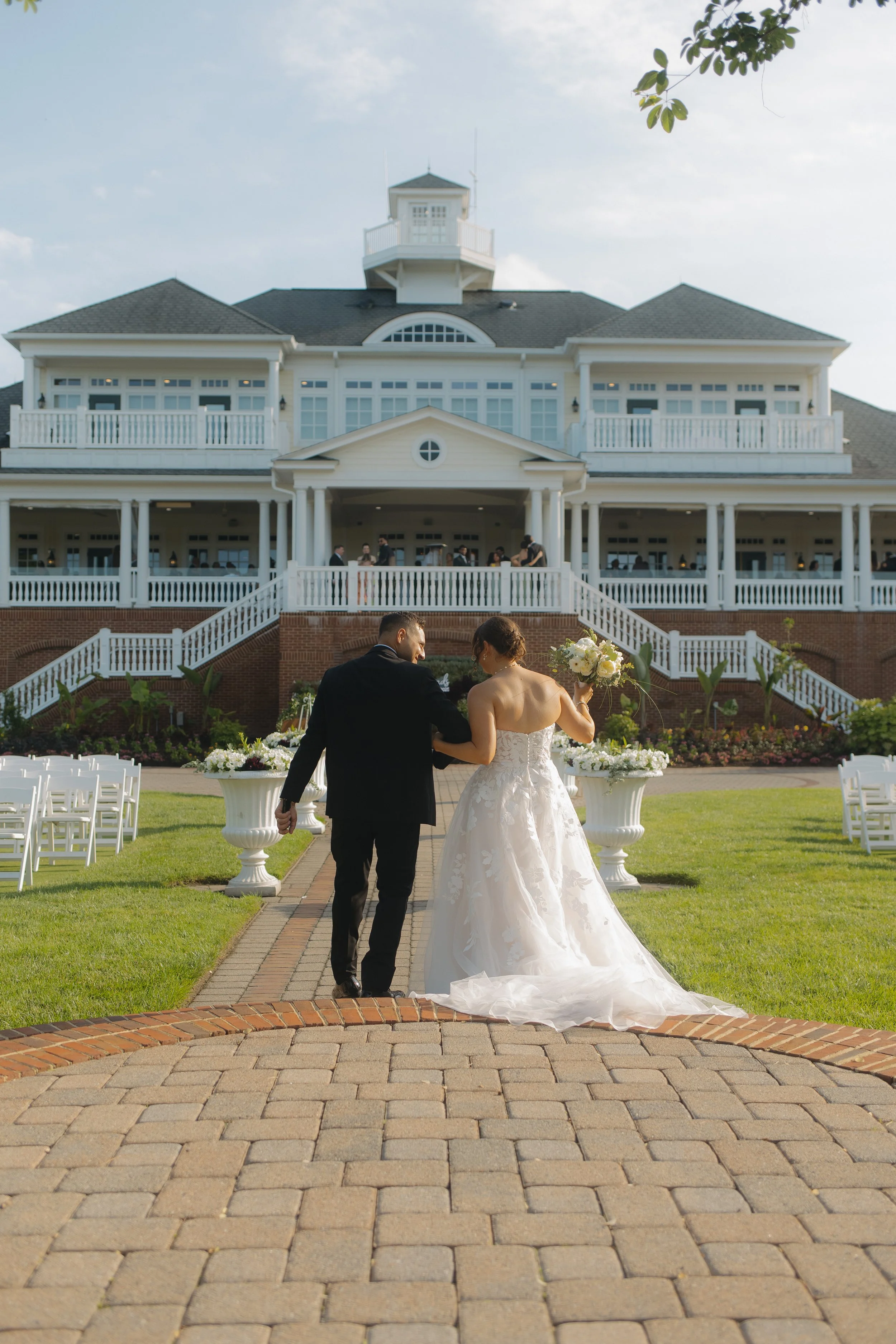 Bride and groom walking towards a large white mansion after their wedding ceremony, with chairs set up on the lawn.