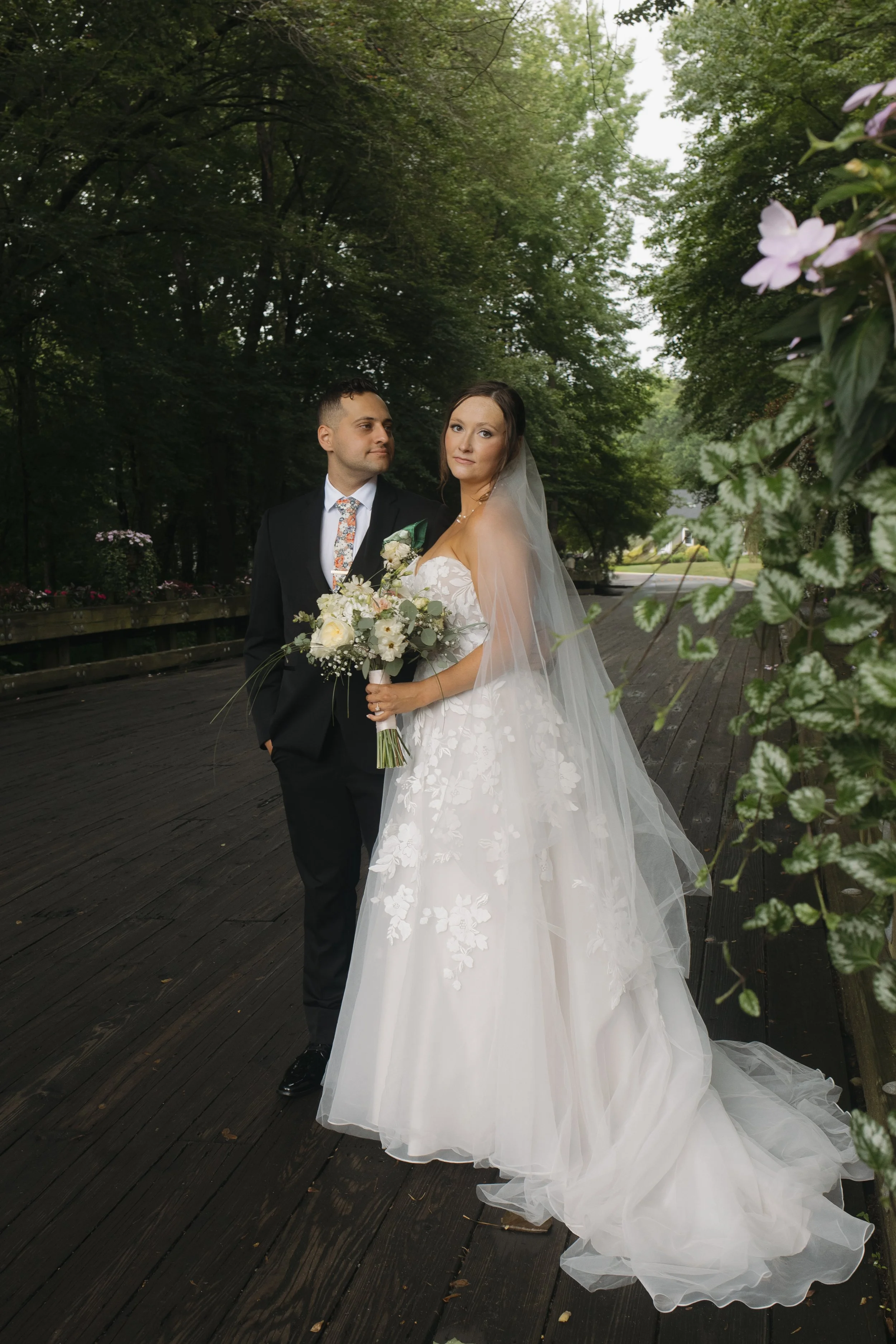 A bride and groom standing on a wooden bridge outdoors during daytime, surrounded by green trees, with the bride holding a bouquet of white flowers and wearing a white wedding dress with a veil, while the groom is dressed in a black suit with a color