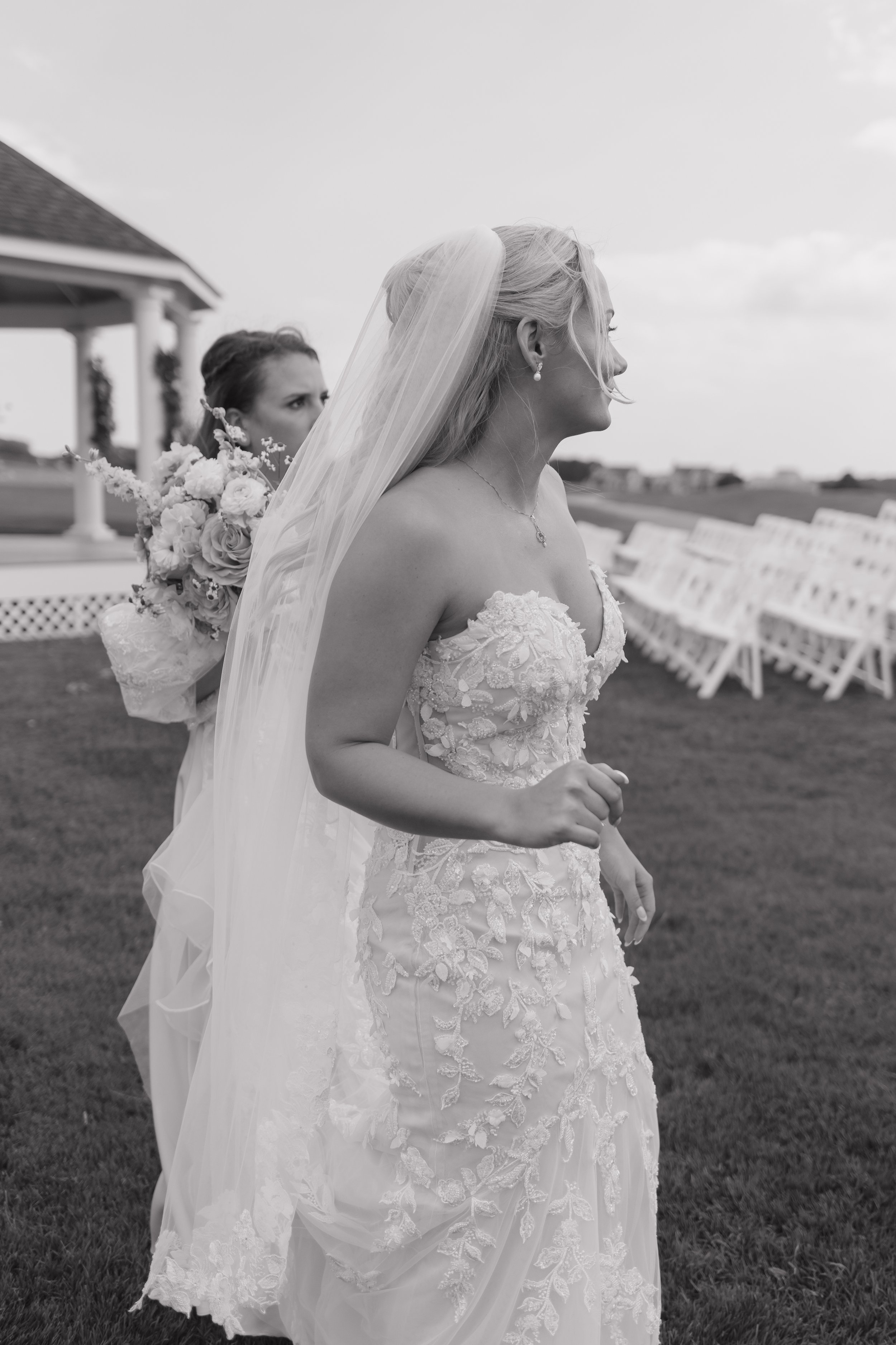 Black and white photograph of two women, one in a wedding dress with a veil, and the other holding a bouquet of flowers, outdoors near a pavilion with white chairs arranged for an event.