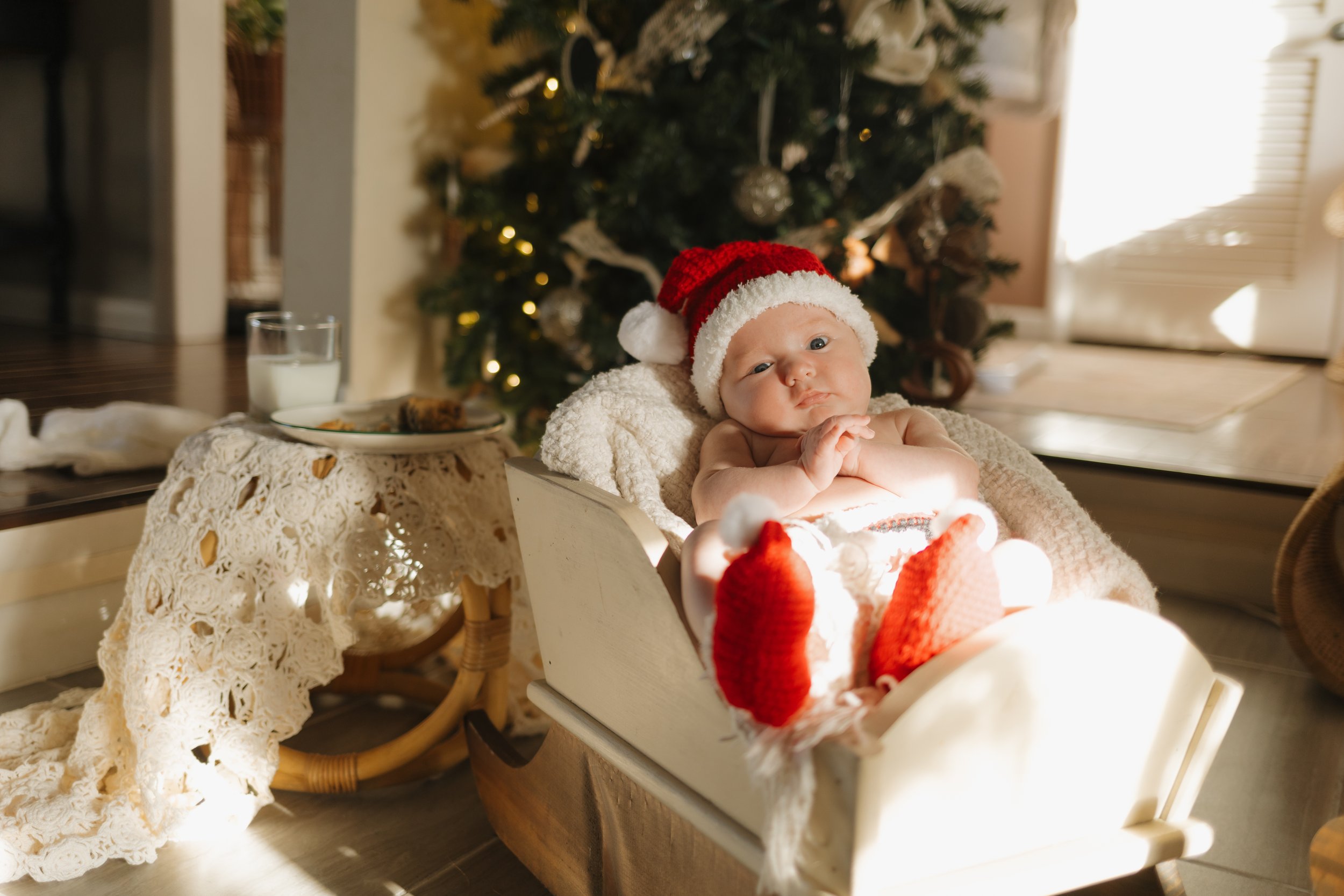 A baby wearing a Santa hat, red socks, and no shirt sitting in a white cradle near a decorated Christmas tree, with a glass of milk, cookie, and lace cloth on a table next to the cradle.