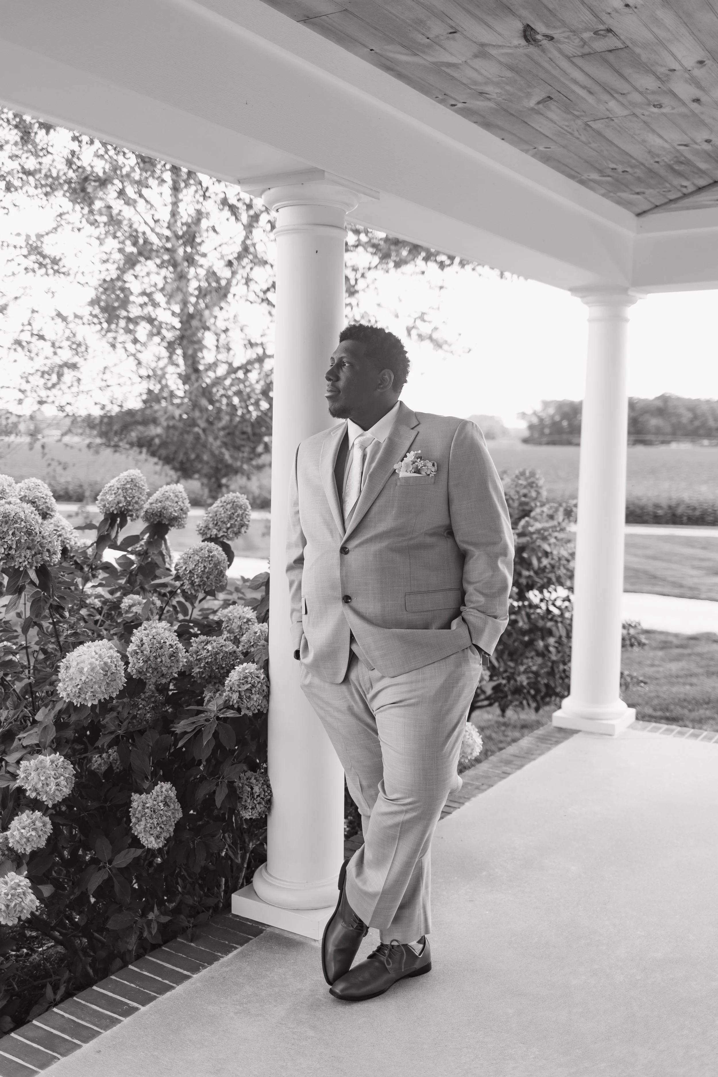 A man in a light-colored suit leaning against a white column on a porch, with hands in pockets, looking to the left, outdoors with trees and flowers around.