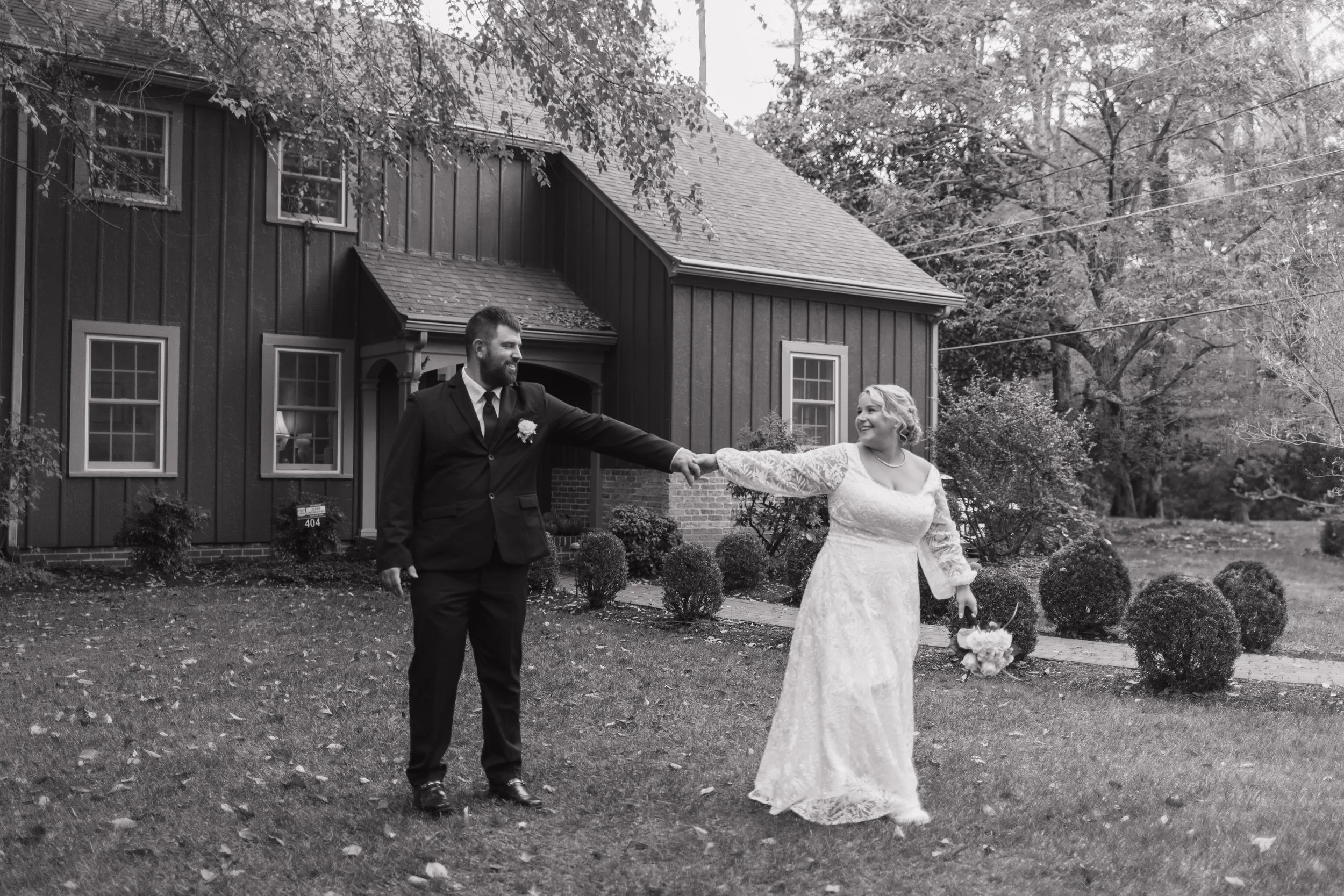 A black-and-white photo of a bride and groom outside a house. The groom, in a suit, is holding the bride's hand as she walks towards him, holding a bouquet. The house has dark vertical panels, multiple windows, and a small porch. There are trees and bushes in the background, and the ground is scattered with fallen leaves.