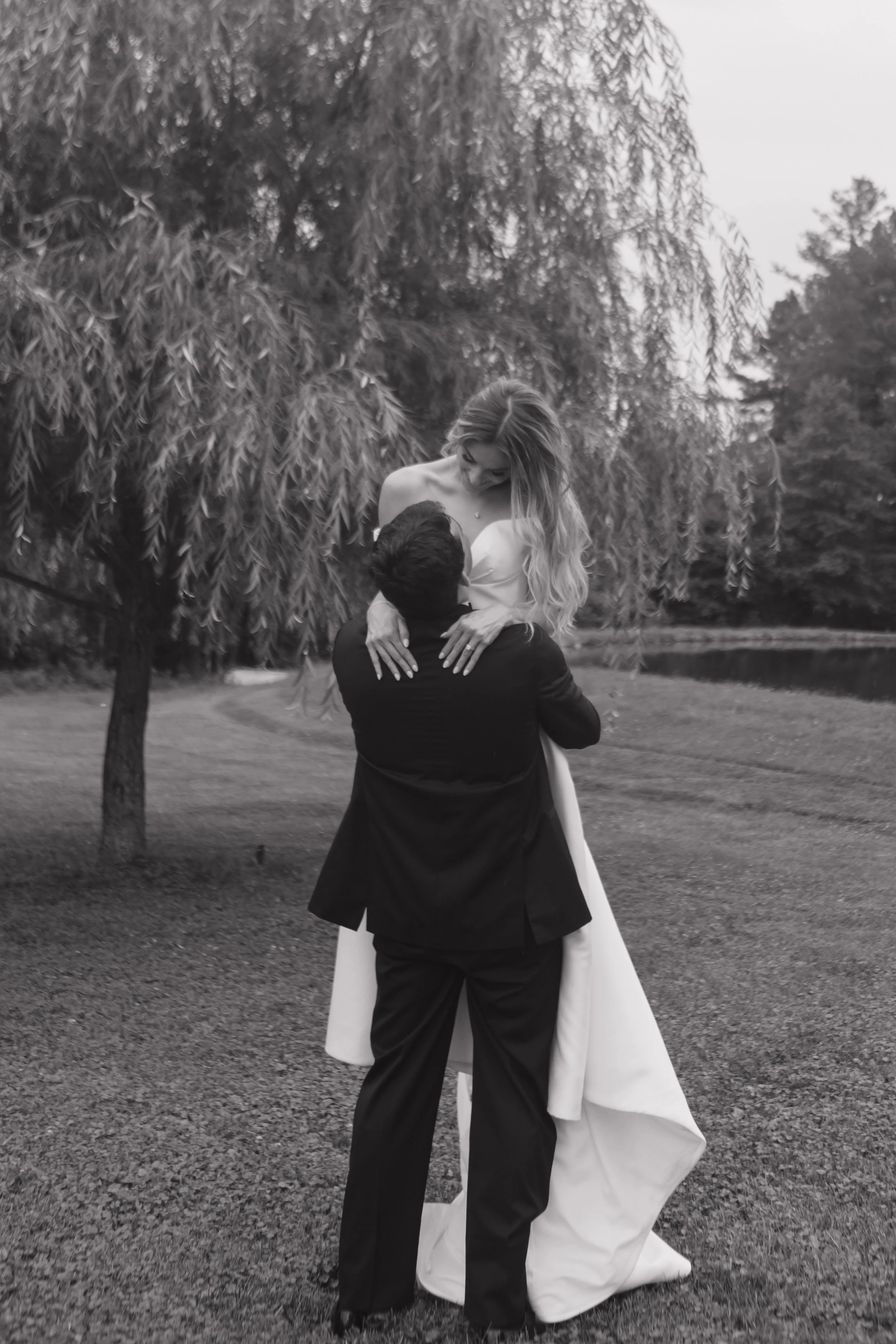 A black and white photo of a bride and groom outdoors near a lake, with the groom lifting the bride in his arms.