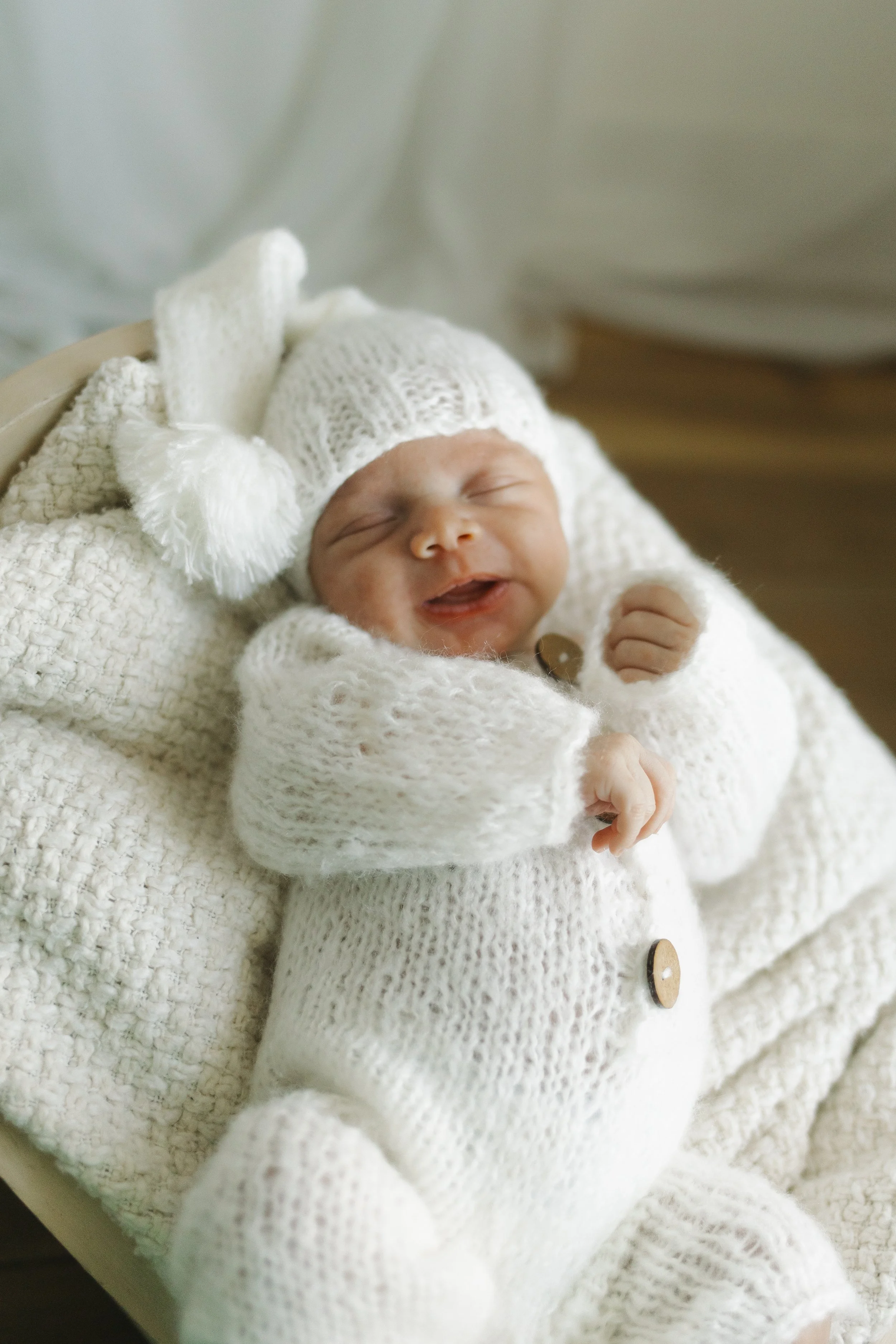 A smiling baby wearing a white knit hat with a pom-pom and a matching white knit onesie, lying on a soft blanket.