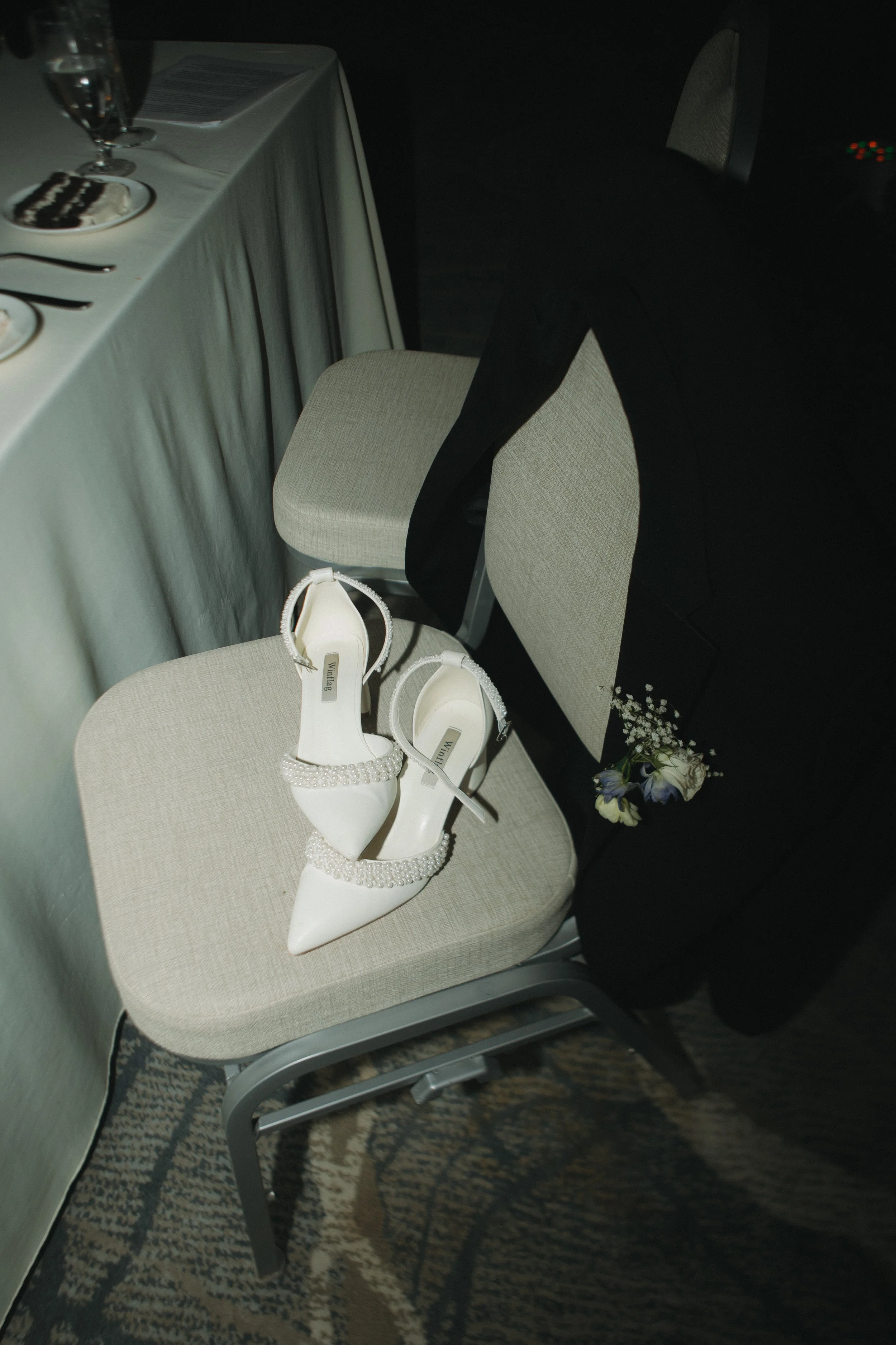 A pair of white wedding high-heeled shoes with pearl embellishments on the straps, placed on a beige chair at a wedding reception table. In the background, there is a black blazer with a small bouquet of flowers tucked into the sleeve.