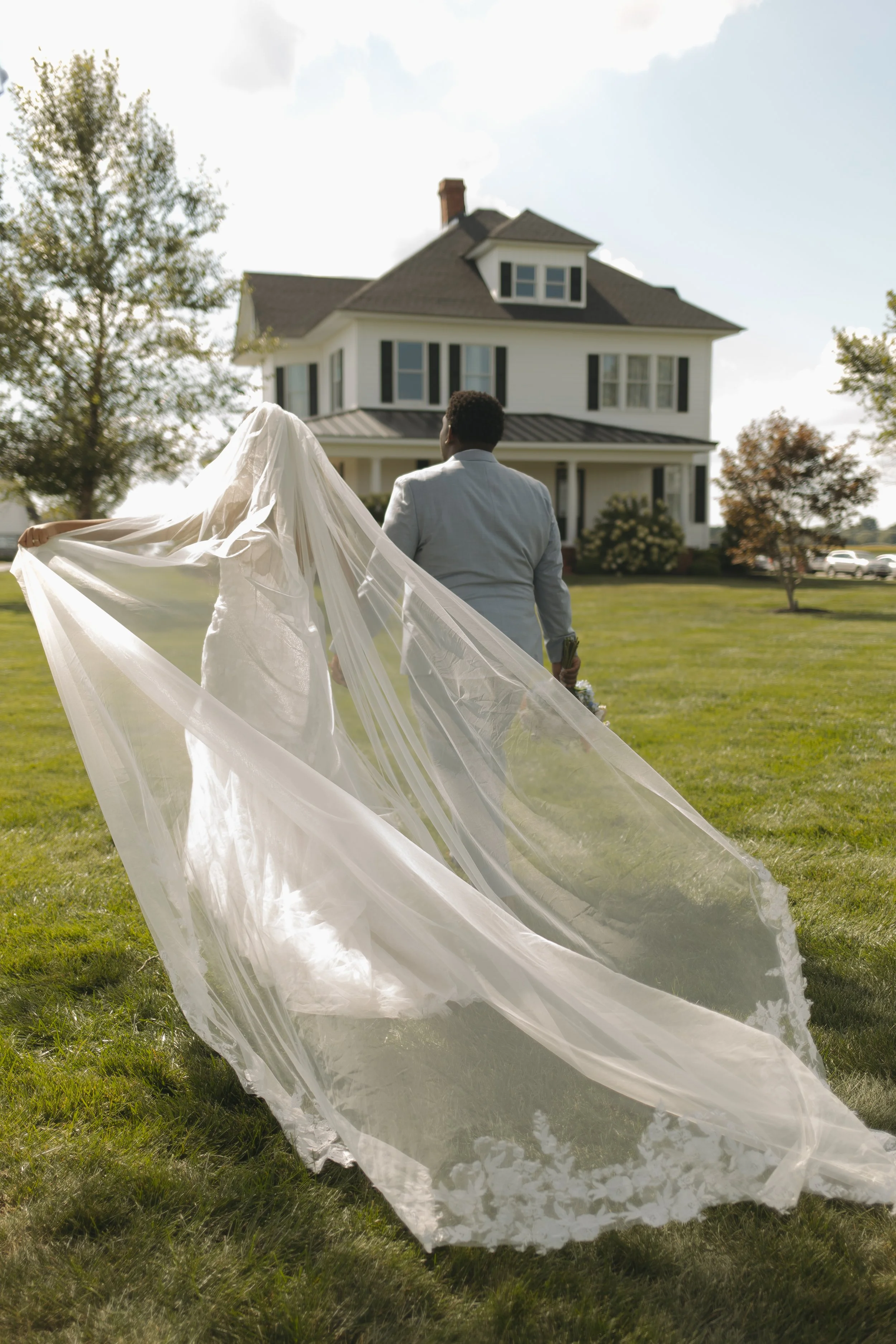 Bride and groom walking toward a large white house on a grassy lawn, with the bride holding her long wedding dress and veil flowing behind her.