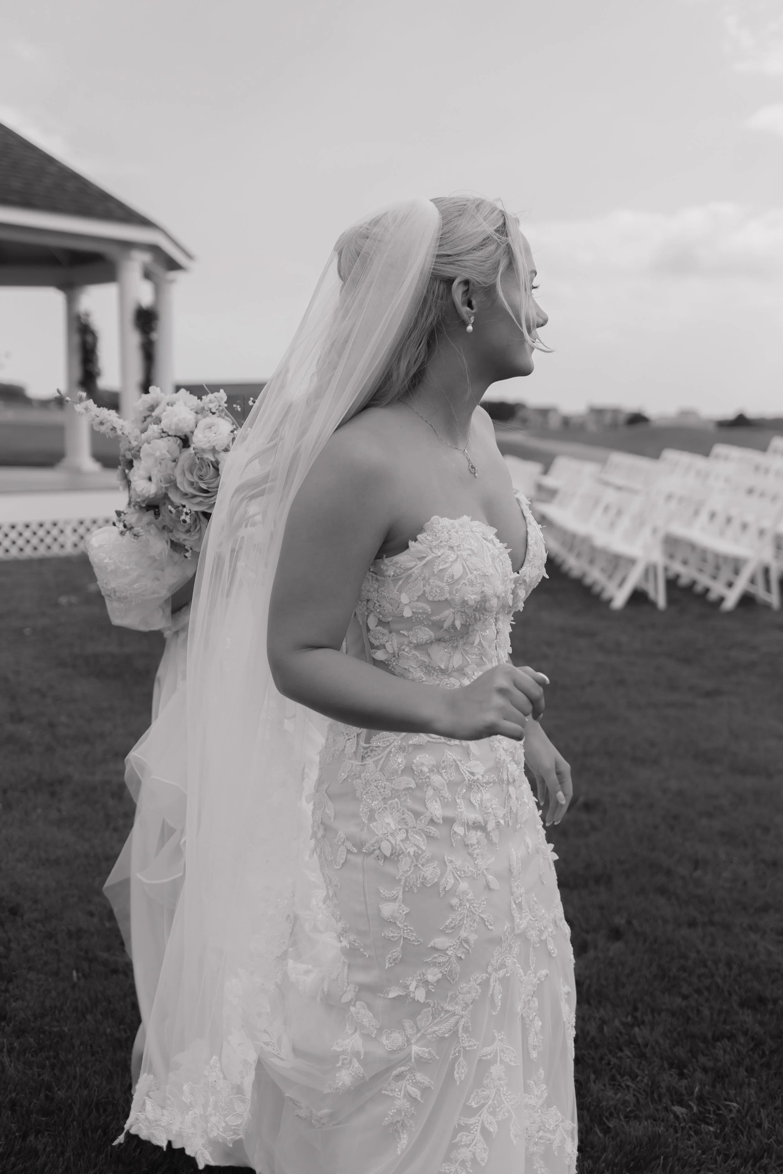 Bride in a strapless lace wedding dress with a veil, holding a bouquet of flowers, walking outside at a wedding venue with chairs and a gazebo in the background.