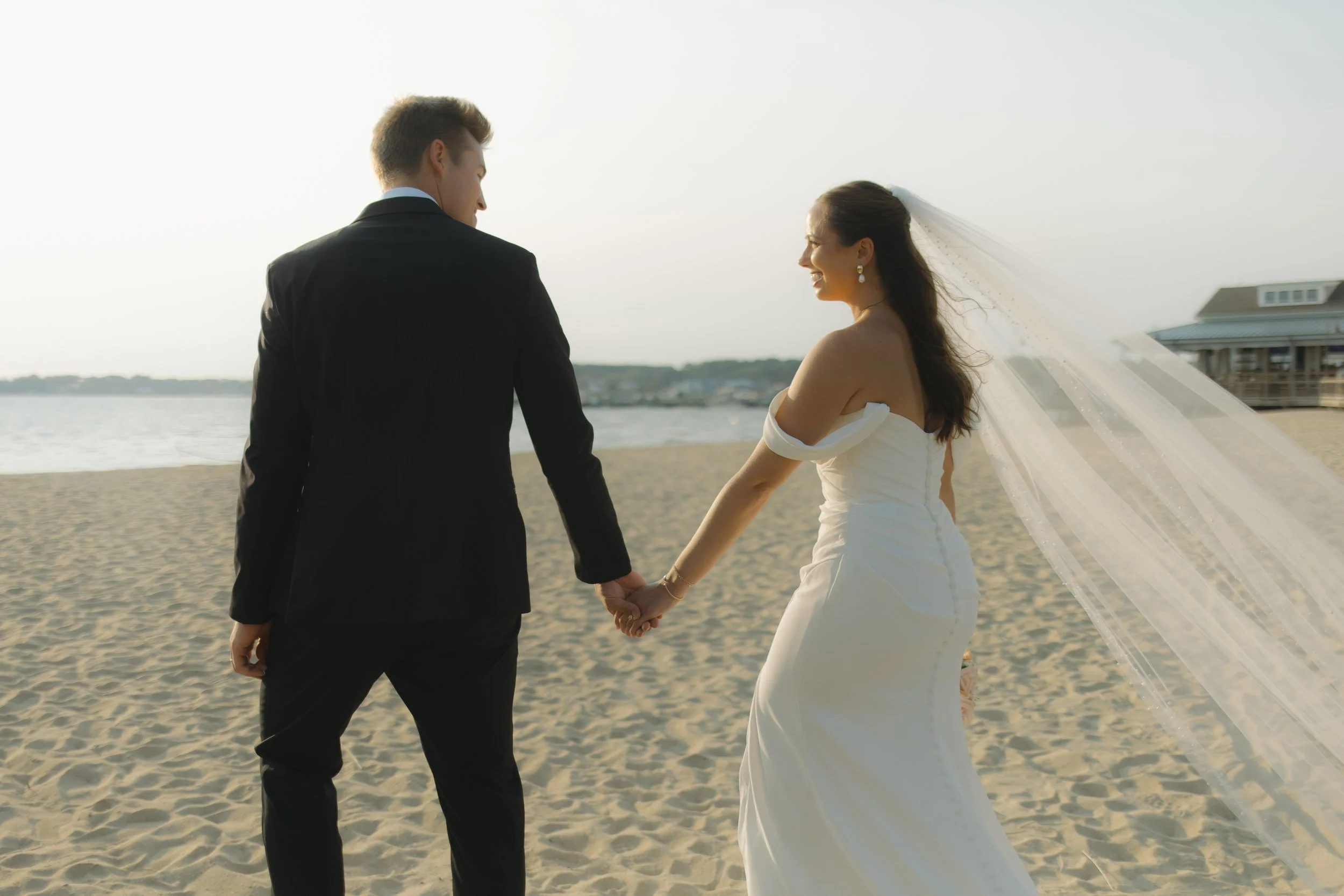 A newlywed couple standing on a beach holding hands, with the bride in a white wedding dress and veil, and the groom in a black suit, facing each other.