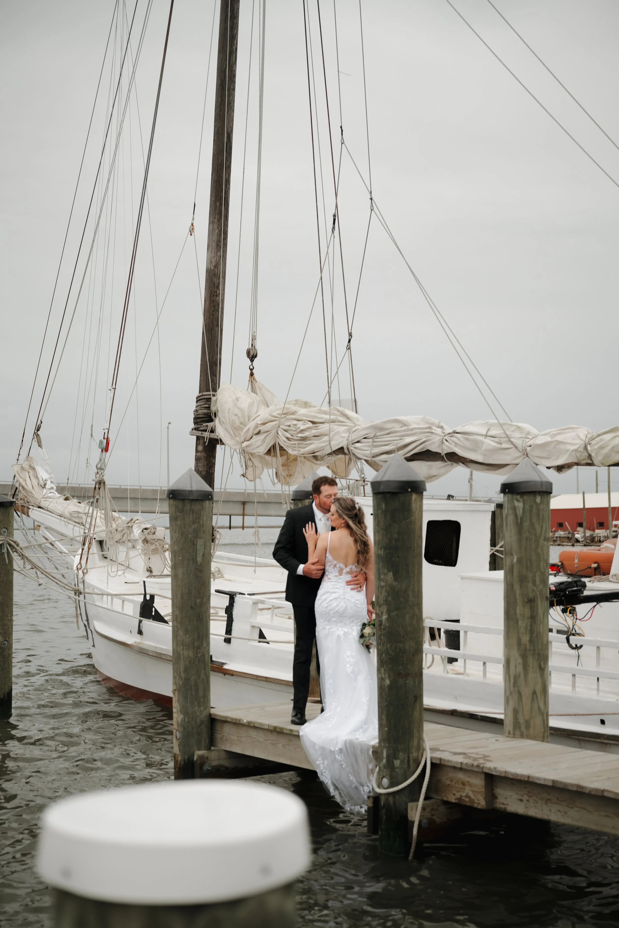 A bride and groom standing on a dock by a sailboat, embracing and gazing at each other, with a gray overcast sky in the background.