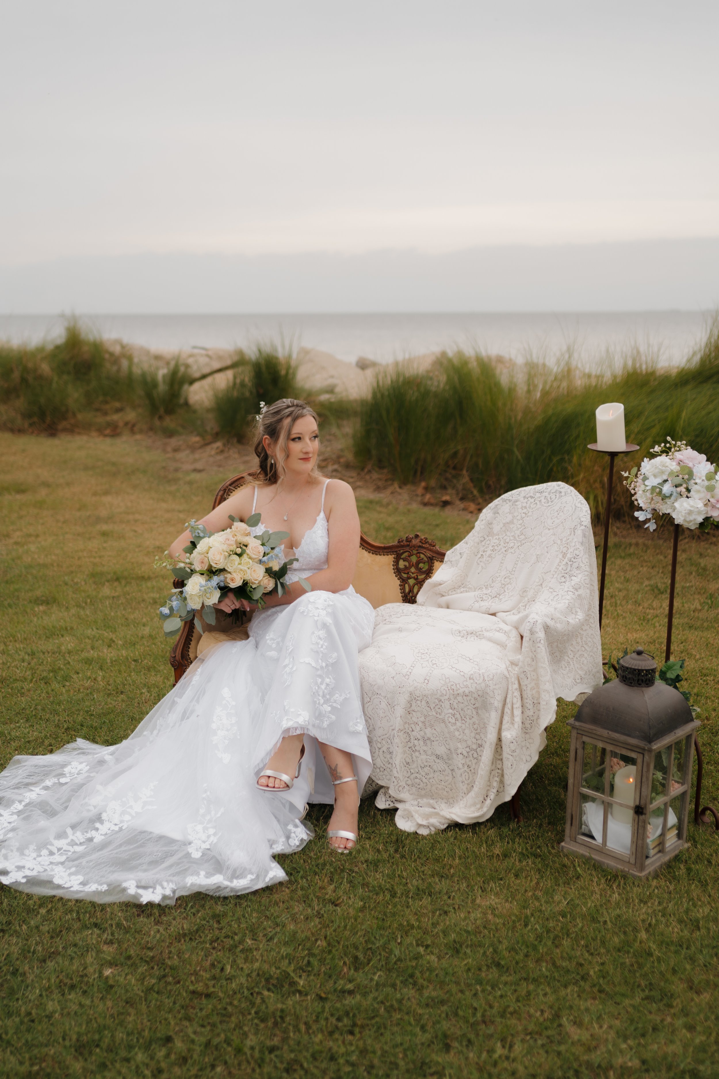 A bride in a white wedding dress sits on a vintage sofa outdoors near the ocean, holding a bouquet of flowers, with candles and floral arrangements nearby.