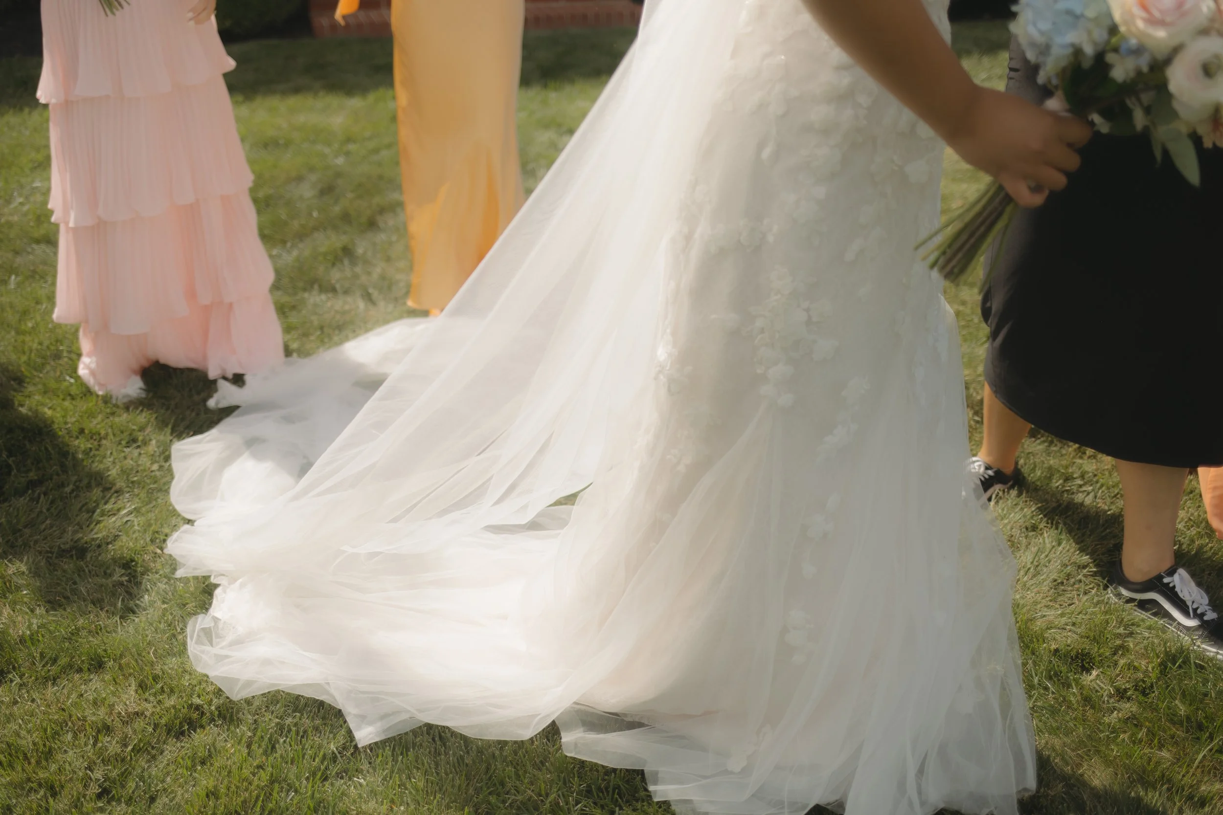 Part of a wedding scene showing the train of a white wedding dress, two women standing nearby, one holding a bouquet, on a grassy outdoor setting.