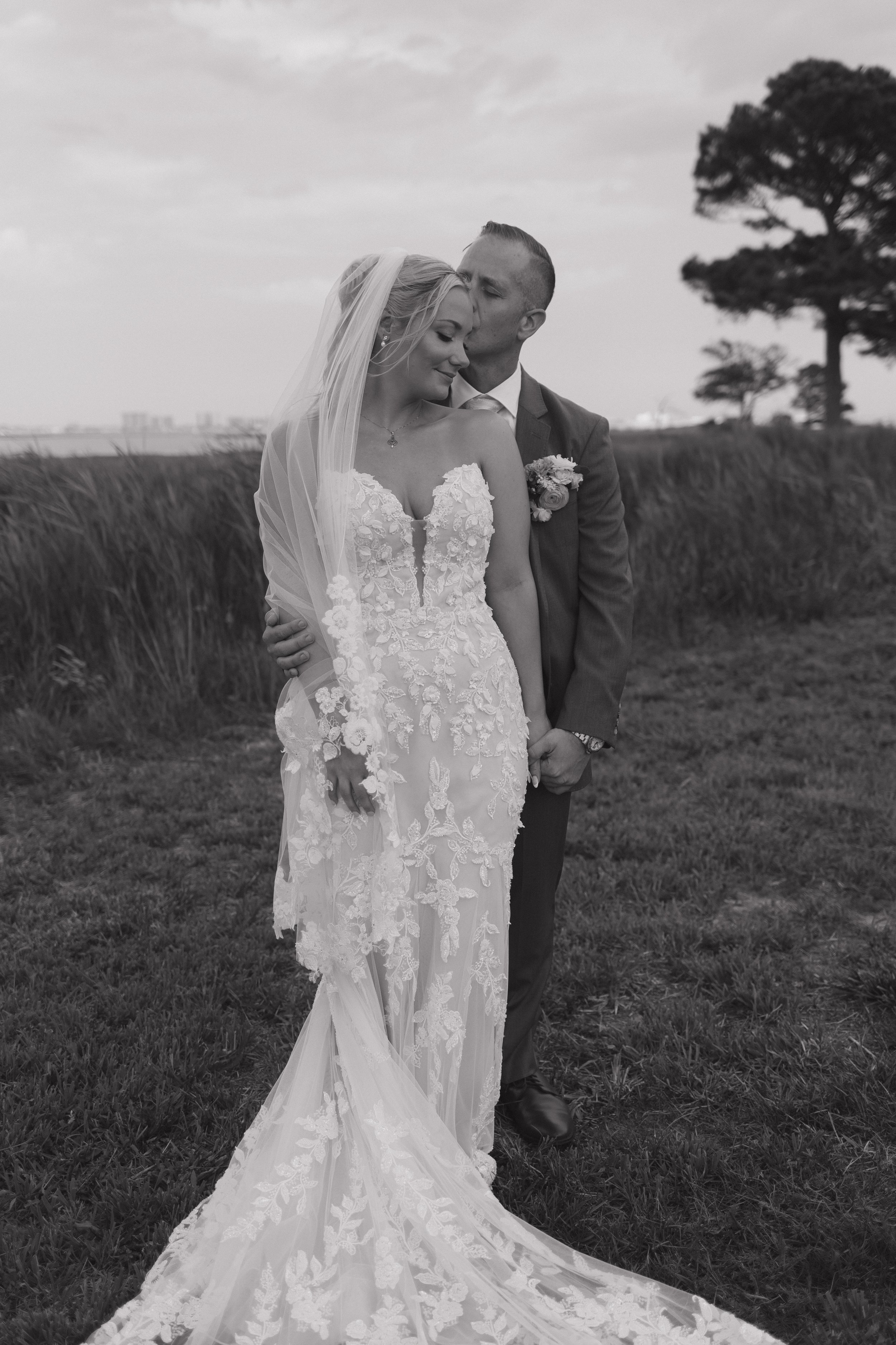 A black and white photo of a bride in a lace wedding gown and a groom in a suit, standing outdoors on grass with a large tree and cloudy sky in the background. The groom is kissing the bride on her forehead, and they are holding hands.