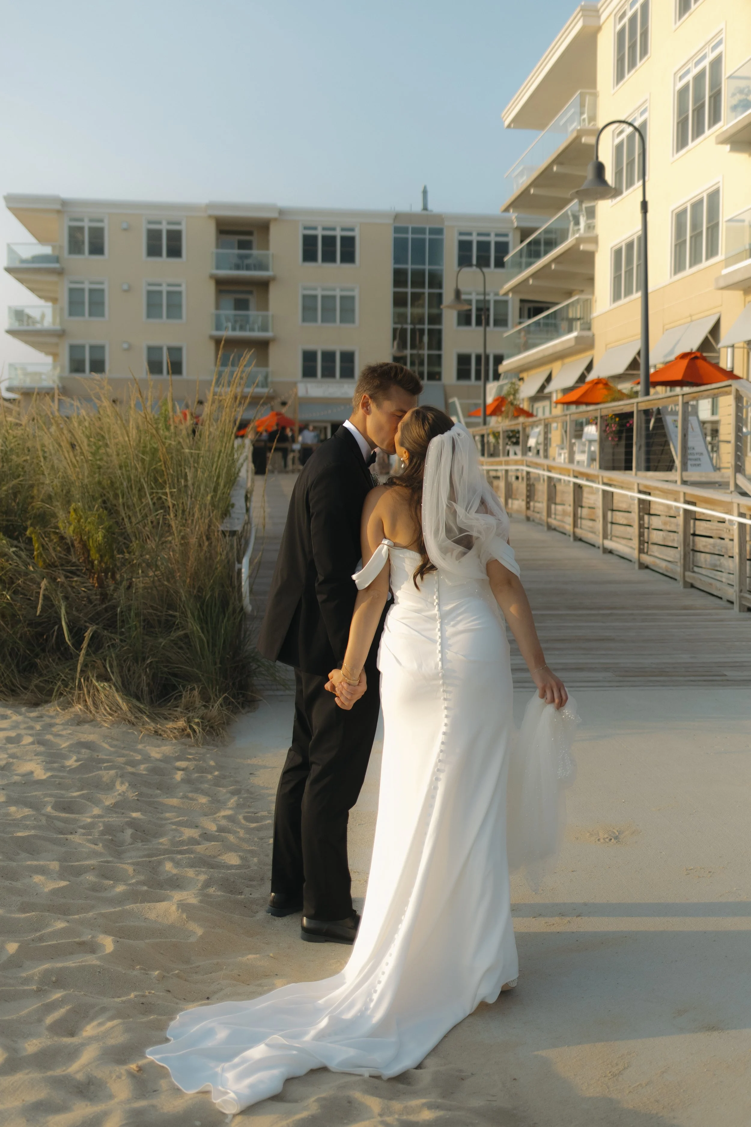 A bride and groom kissing on a sandy beach near a wooden walkway with a modern apartment building in the background.