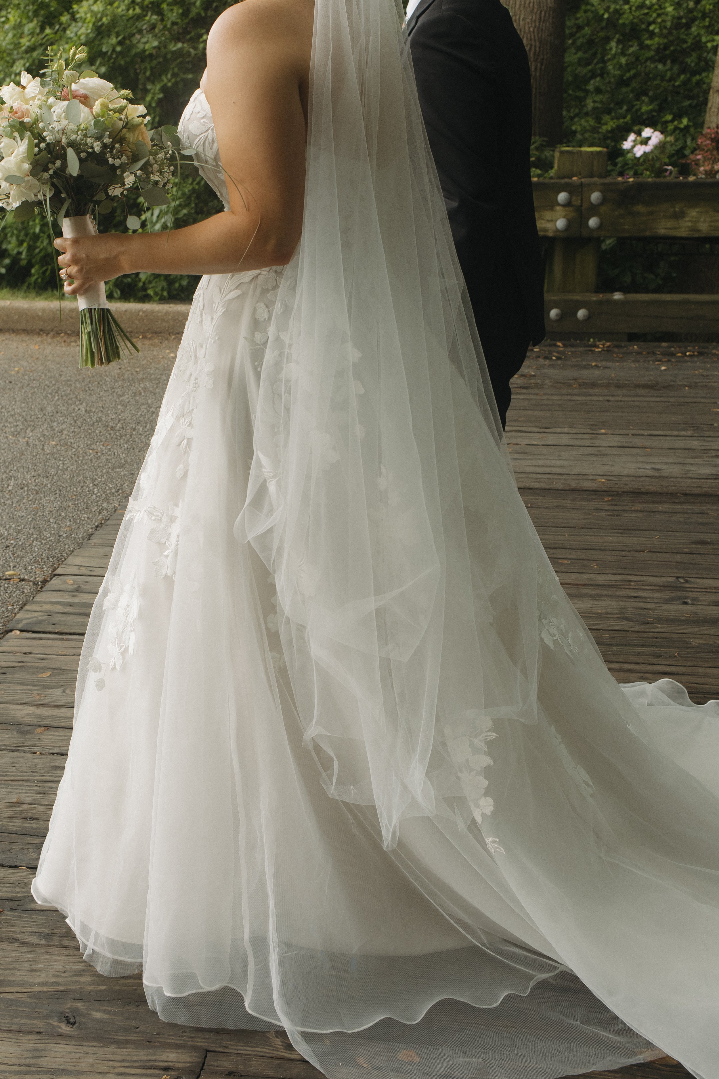 Close-up of a bride holding a bouquet of flowers and a man in a suit standing next to her, on a wooden outdoor pathway surrounded by greenery.