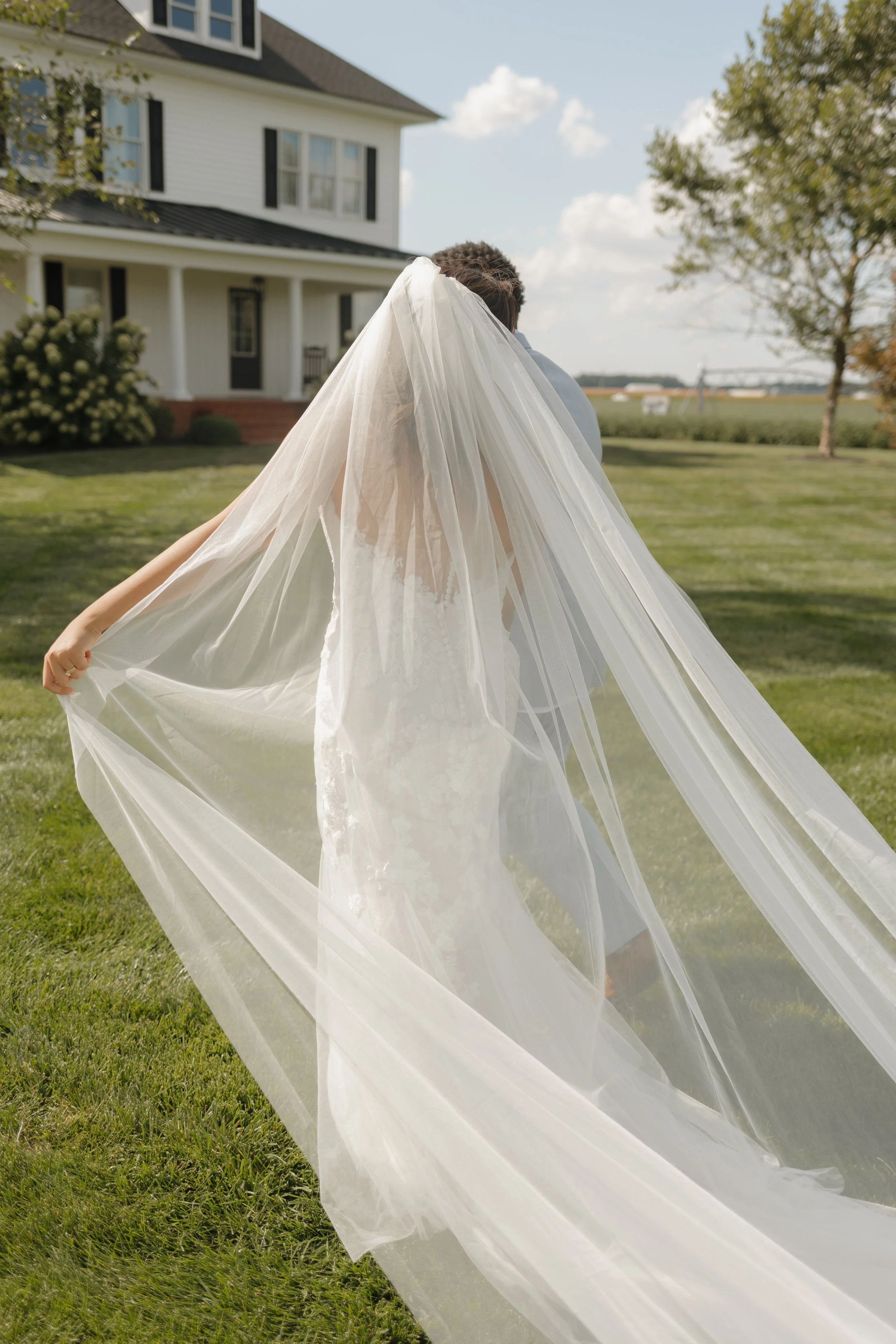 Bride in a wedding dress and veil holding the edges of her long veil, standing on a lawn with a white house and trees in the background.