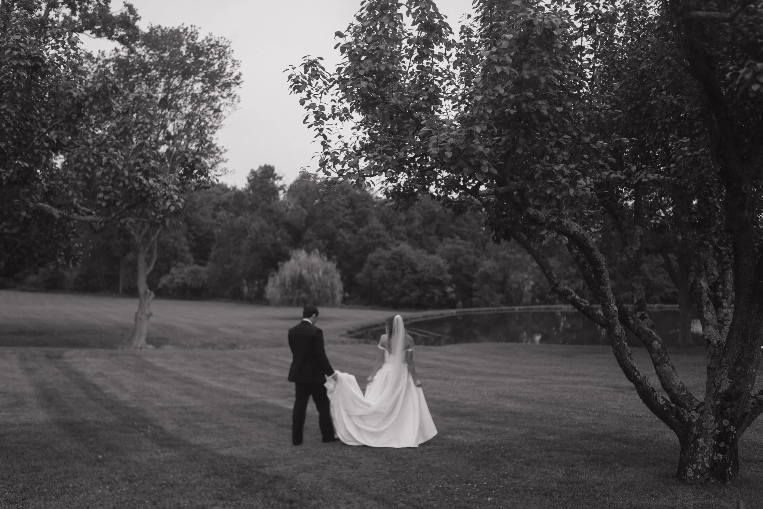 A black and white photo of a bride and groom walking hand in hand in a park, with large trees and a pond in the background.