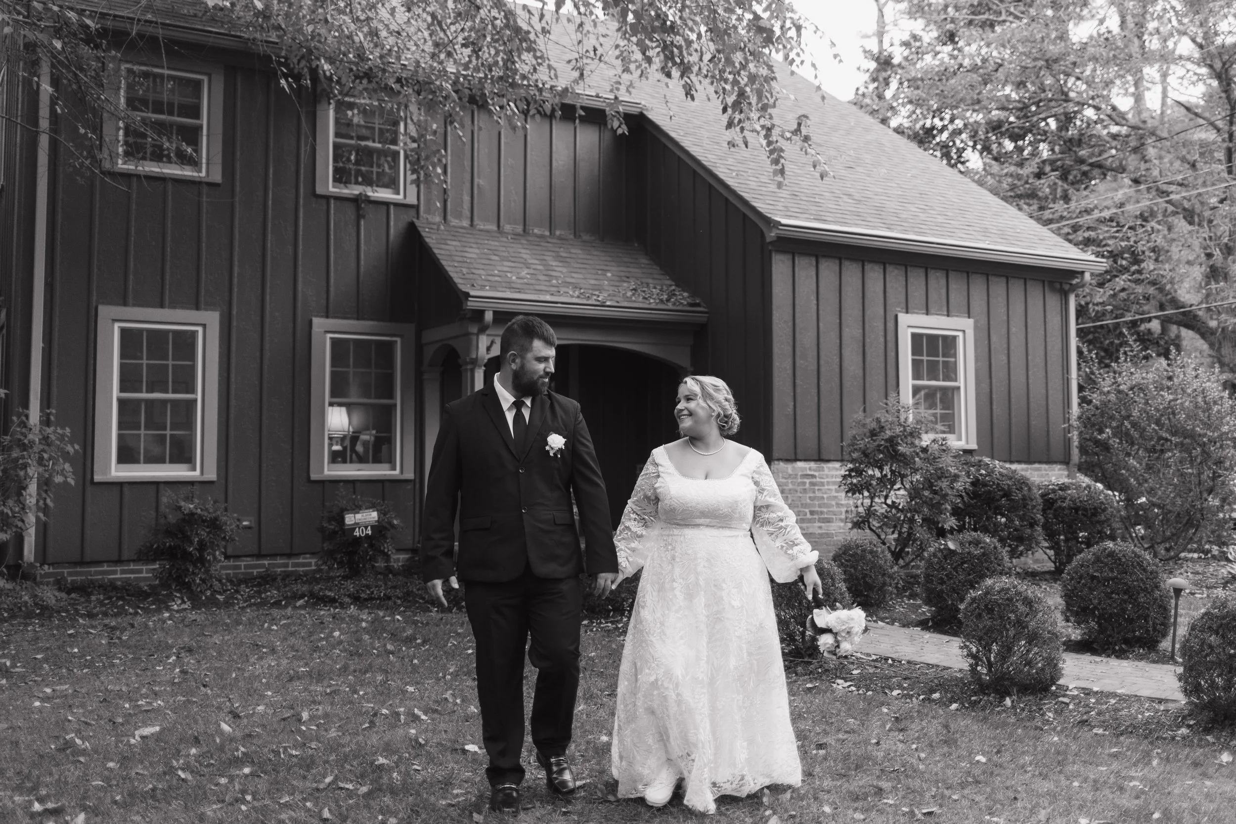 A bride and groom walking hand in hand outside a house, smiling at each other, on their wedding day in black and white.