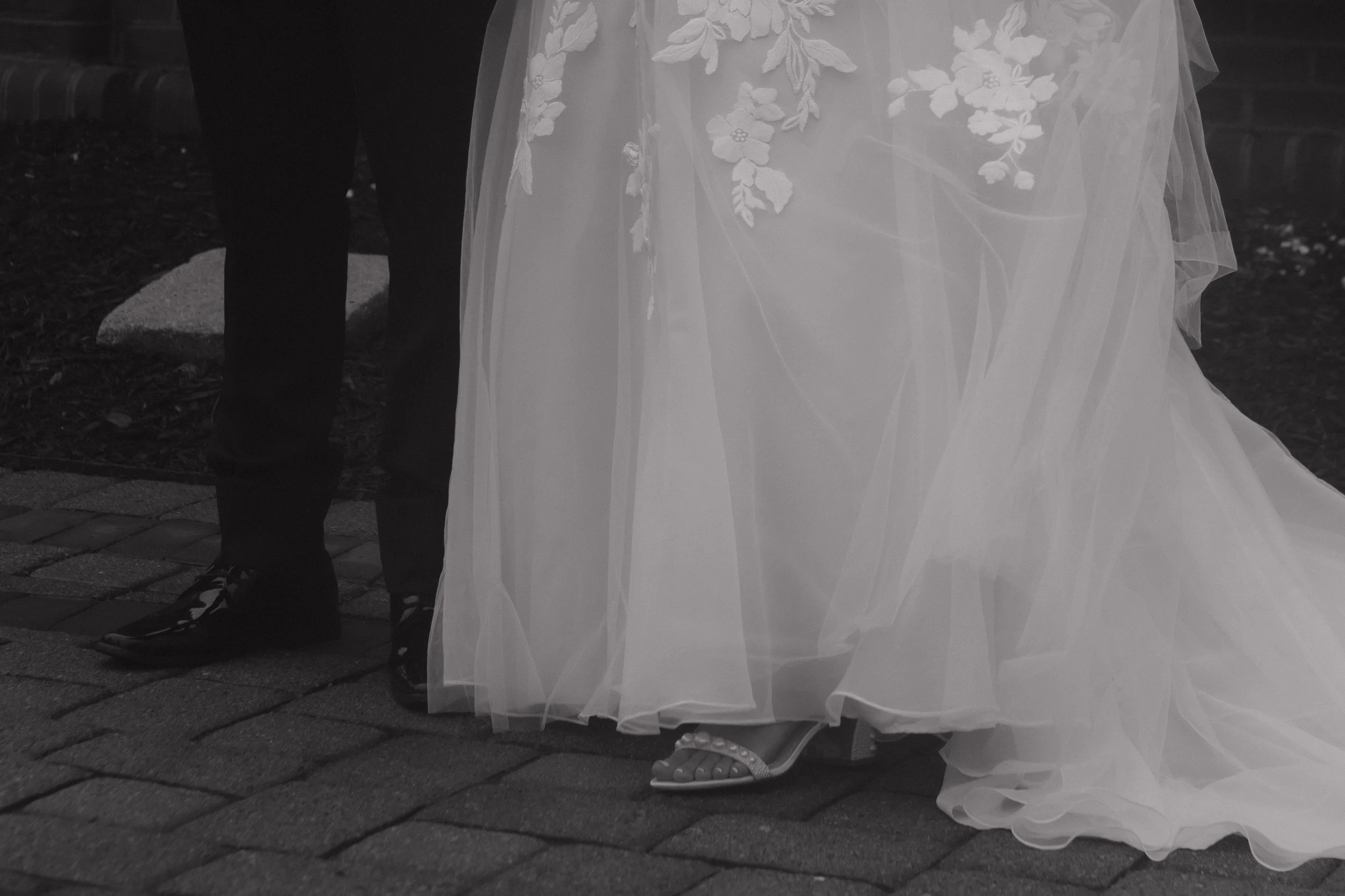 Close-up of a bride in a wedding dress and high heels standing next to a groom in dark trousers and shoes, on a brick pathway.