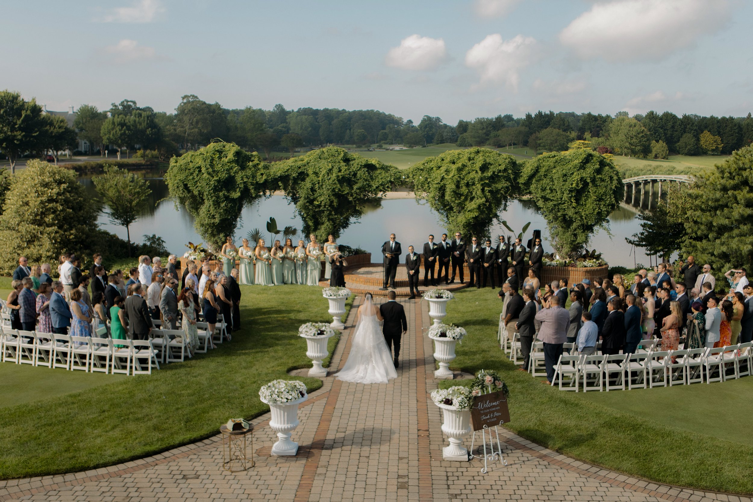Wedding ceremony outdoors by a lake with guests seated on white chairs, bride and groom walking down the aisle, bridal party standing at the altar, green trees, cloudy sky, and water in the background. Country club ceremony, timeless photography, 