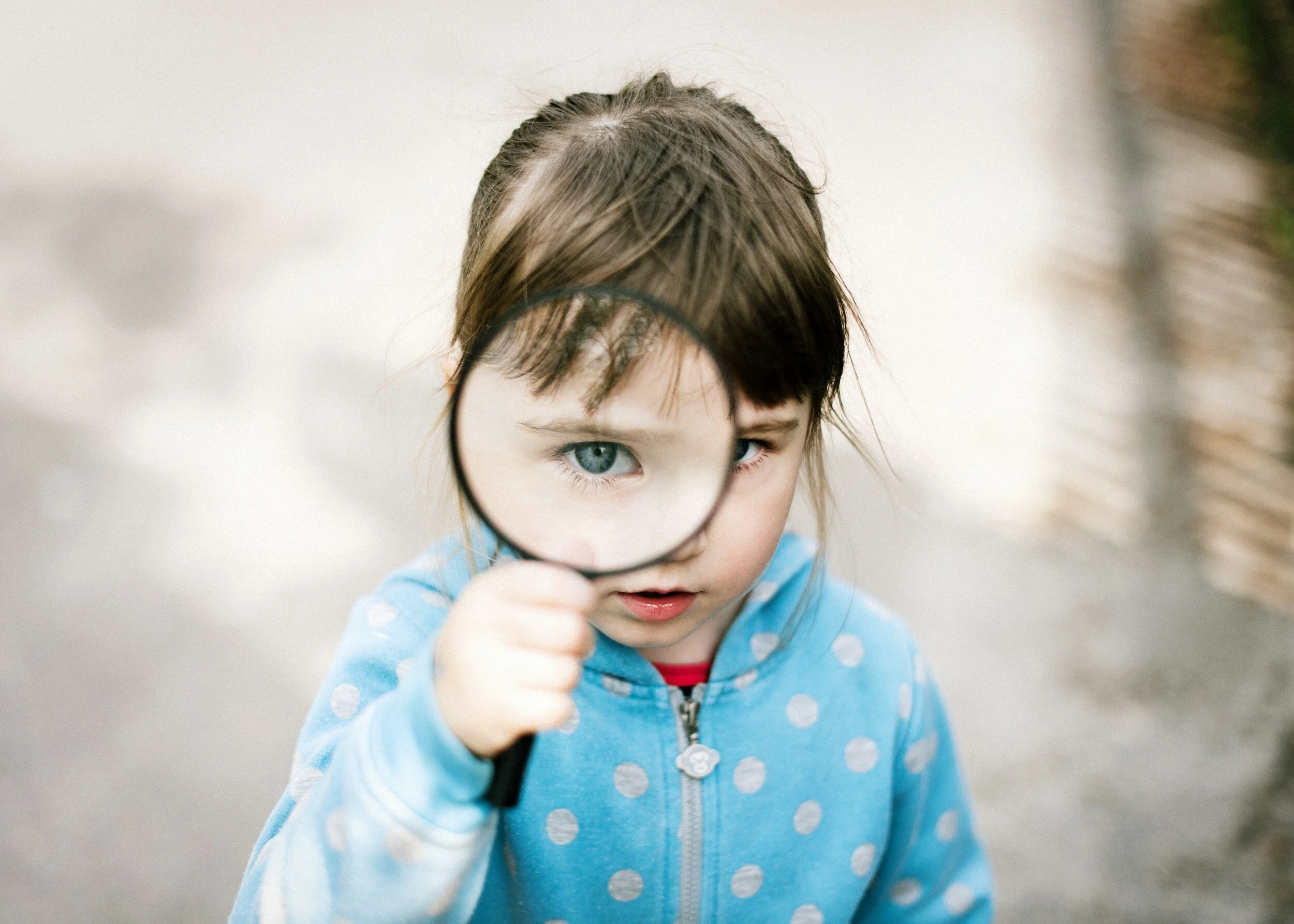 Child holding a magnifying glass, wearing a blue polka dot hoodie, looking through the lens.