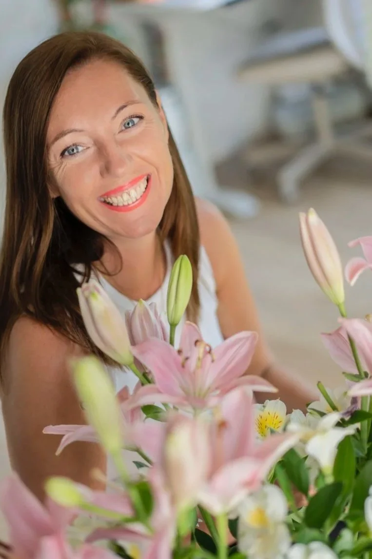A woman with long brown hair smiling at the camera, surrounded by pink lilies and other flowers.