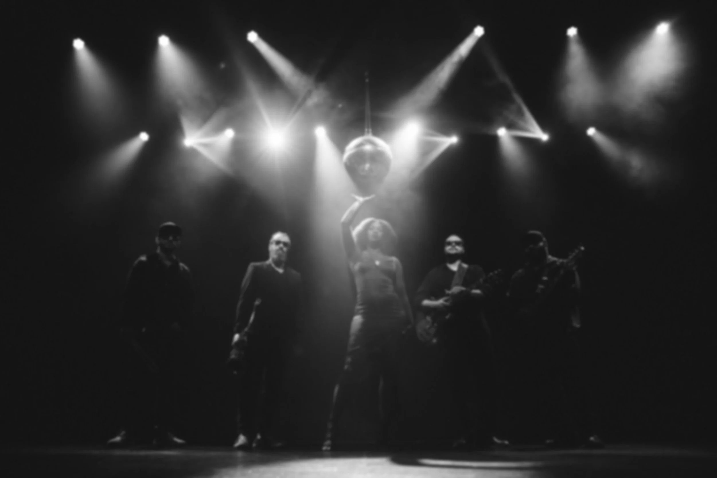 Black and white photo of five performers on stage with a disco ball overhead, stage lights shining down, and one person holding a microphone, possibly singing or speaking.