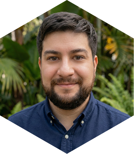 Headshot portrait of software engineer Jesus Gonzalez, a man in his late thirties wearing a navy blue button down shirt in front of a tropical foliage background. 