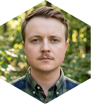 Headshot of Garrett Burr, a young man with short brown hair and blue eyes wearing a blue and green flannel in front of a blurred foliage background