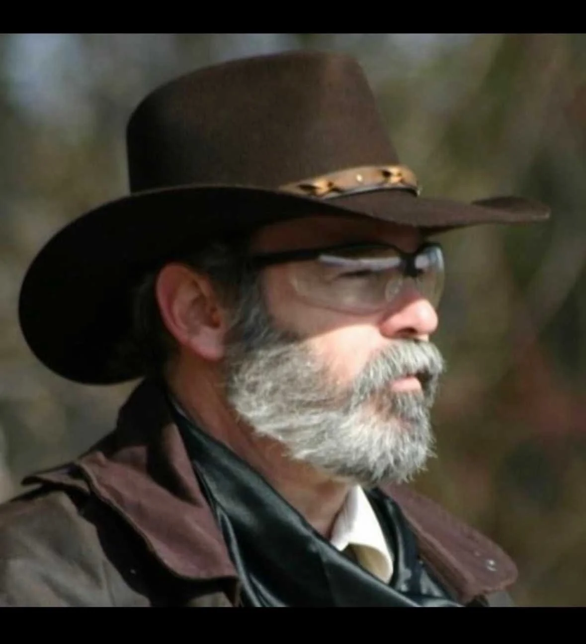 Profile of an older man with gray beard and glasses, wearing a brown cowboy hat with a decorative band, an outdoorsy jacket, and a leather shirt.